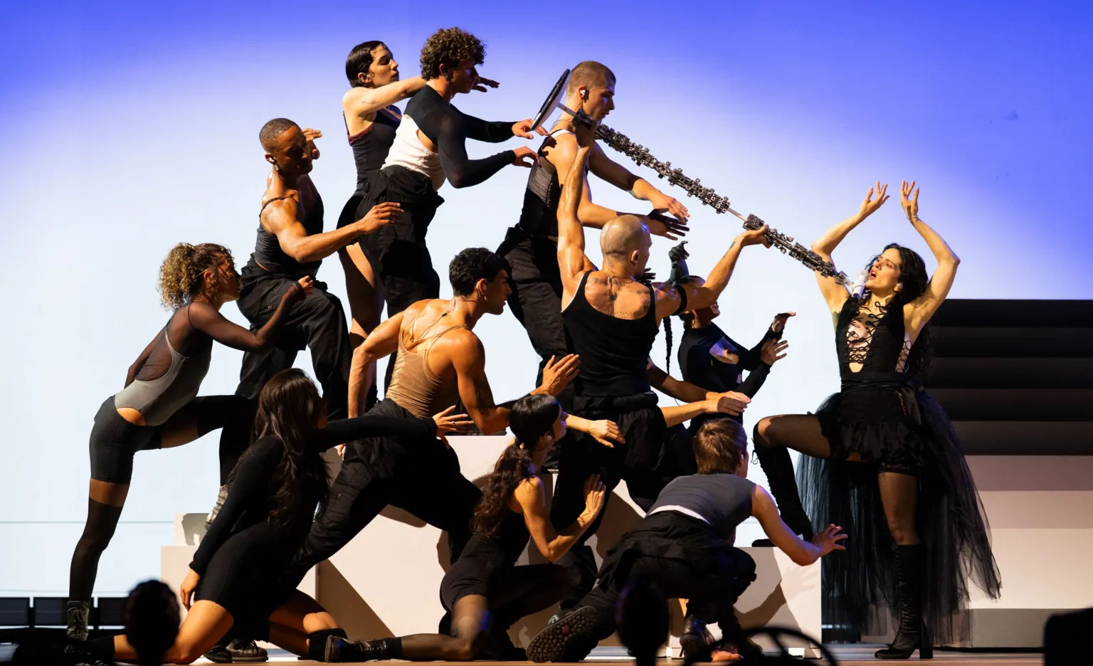 Samir Hussein Rosalia on stage posing with her arms in the air, facing 11 dancers who are pointing a long stick or brush at her
