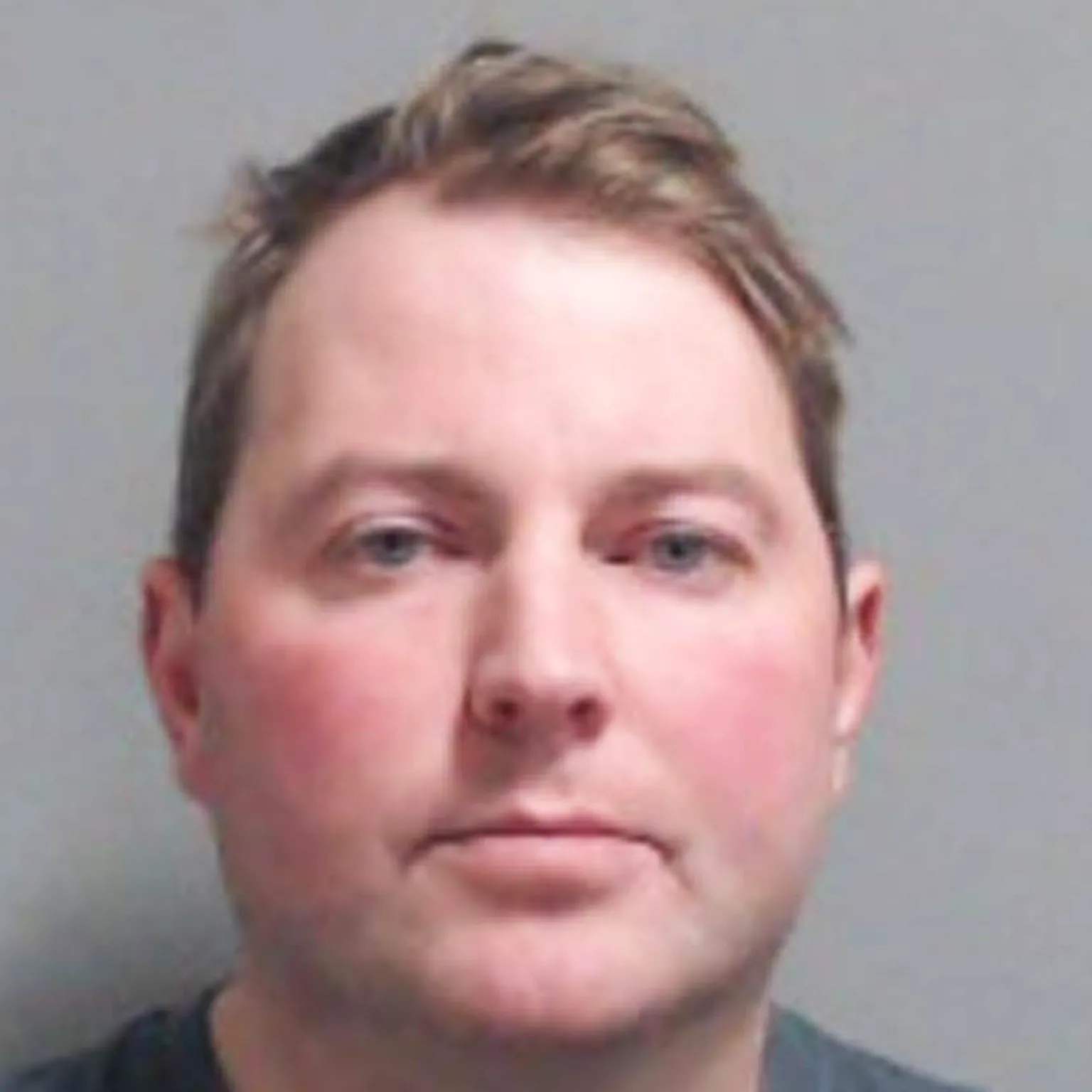 PSNI Jonathan Creswell has short brown hair parted to the side and light blue eyes. He is wearing a blue/grey t-shirt and is staring straight ahead in a police mugshot. 