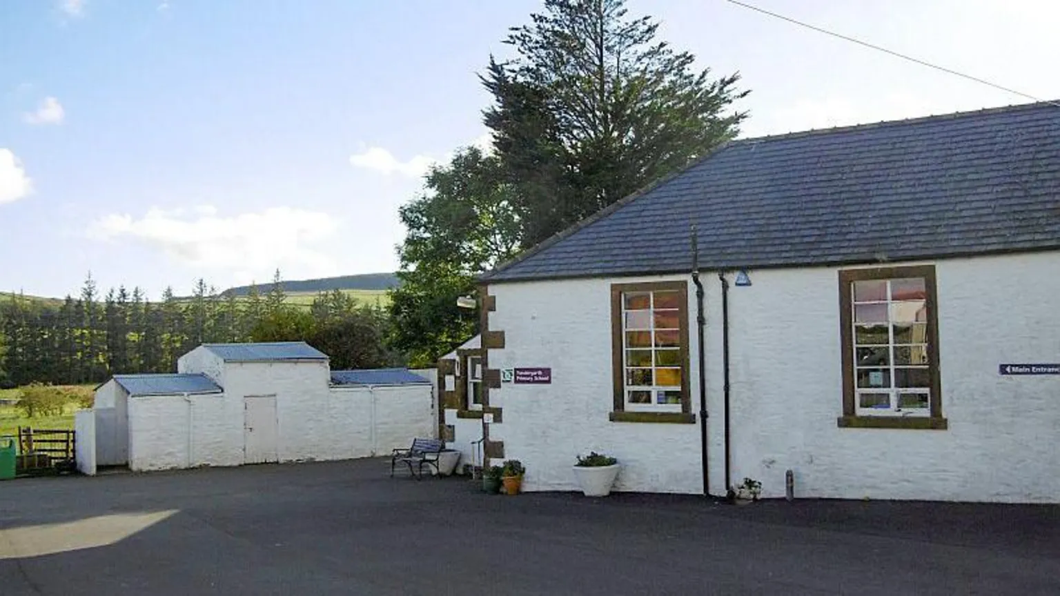 Richard Webb A small whitewashed rural school building, single-storey with white outbuildings and tarmac in front with trees and hills in the distance