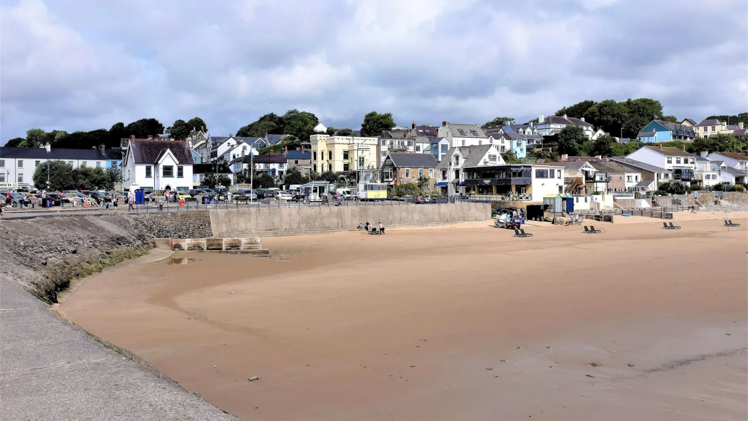 Oscar Hill/ This image shows Saundersfoot beach, the sand is beige and the sky is blue. There's a stone wall separating the sandy beach from a collection of brightly coloured buildings. 