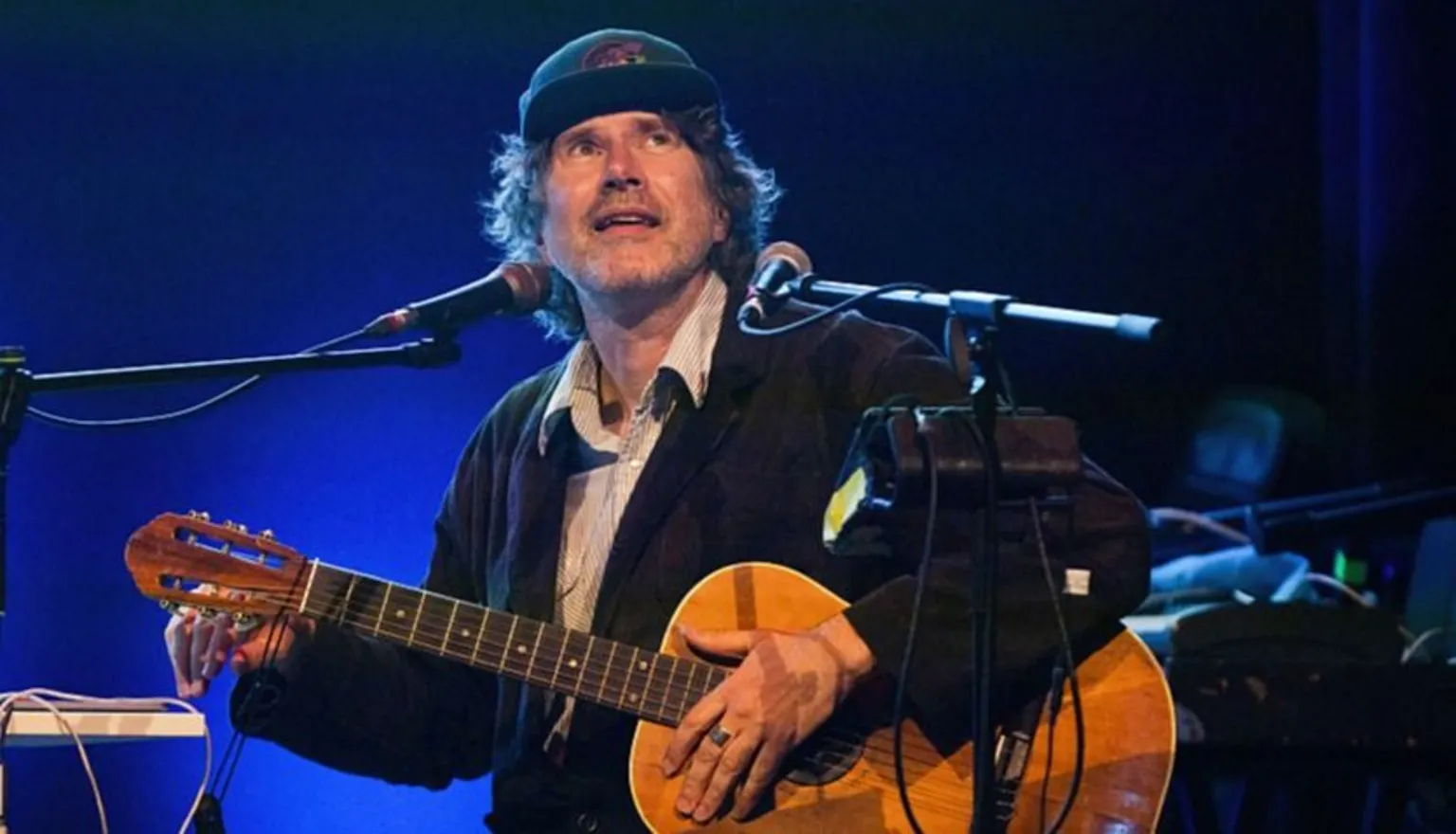  Gruff Rhys, on stage with an acoustic guitar, dressed in a cap and dark jacket, smiling at the crowd.