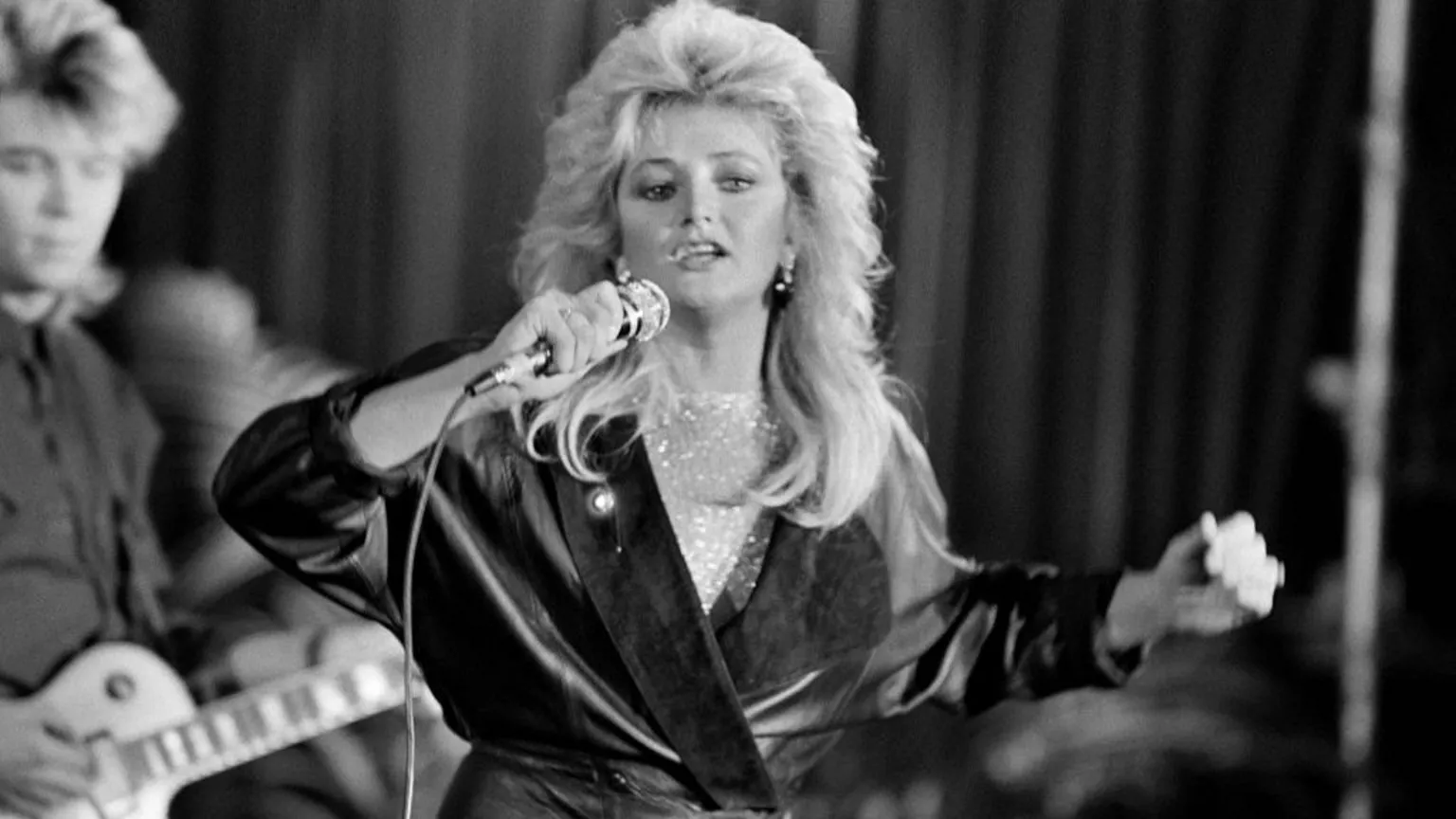  Black and white image of Bonnie Tyler, with big blonde hair, singing into a microphone. She is wearing a leather trench coat, with a musician playing a guitar in the background. 