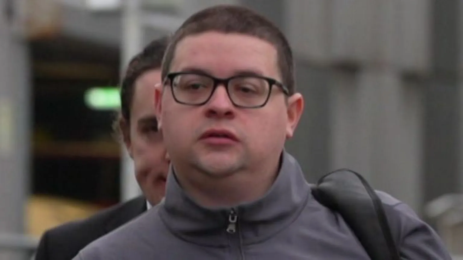Ieuan Crump is wearing a grey turtleneck jacket, black glasses and a black bag strap over his shoulder. 