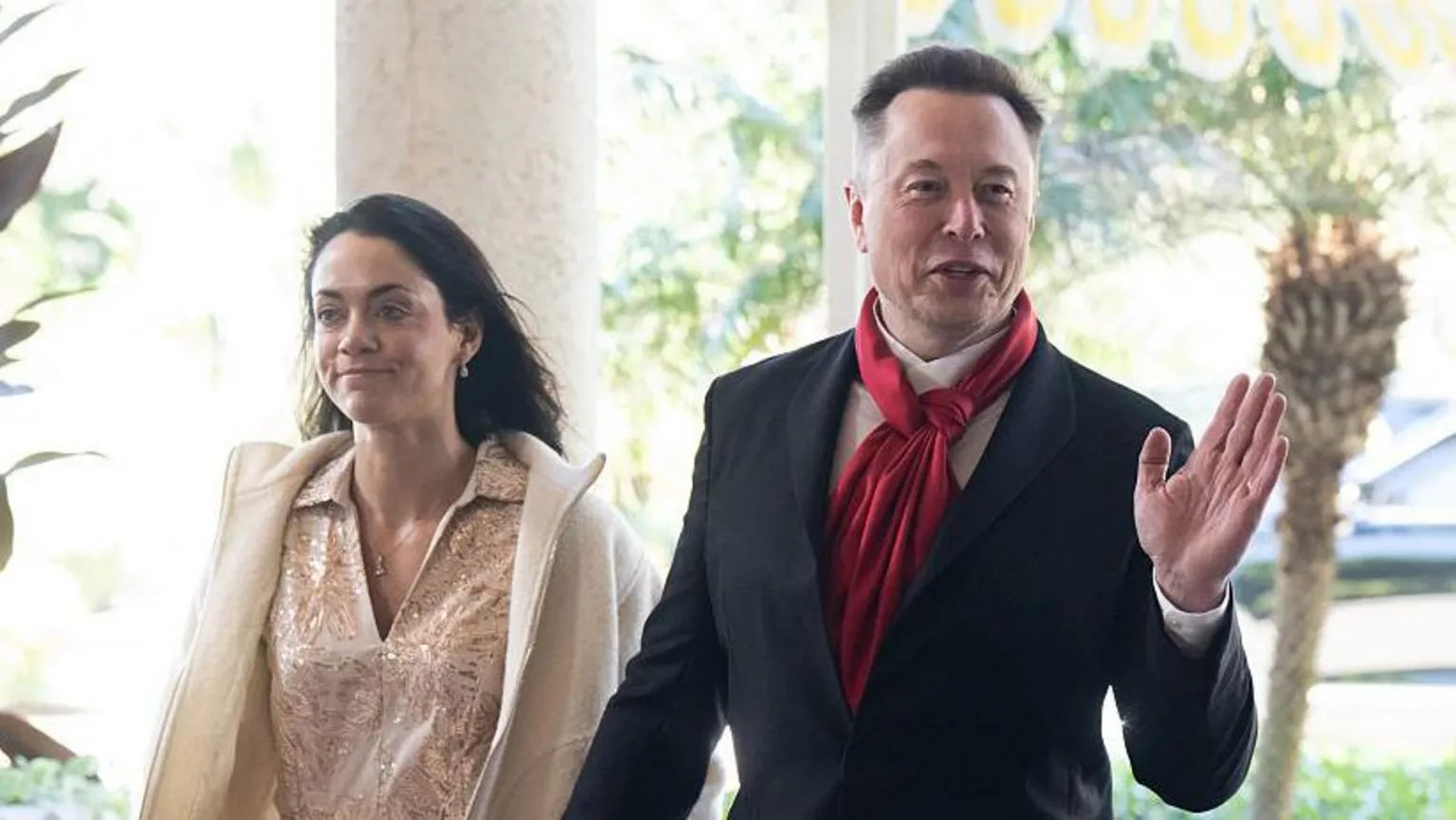 AFP via Shivon Zilis wearing a pale dress and jacket, with Elon Musk wearing a suit and red scarf. Both are smiling and Musk is waving.