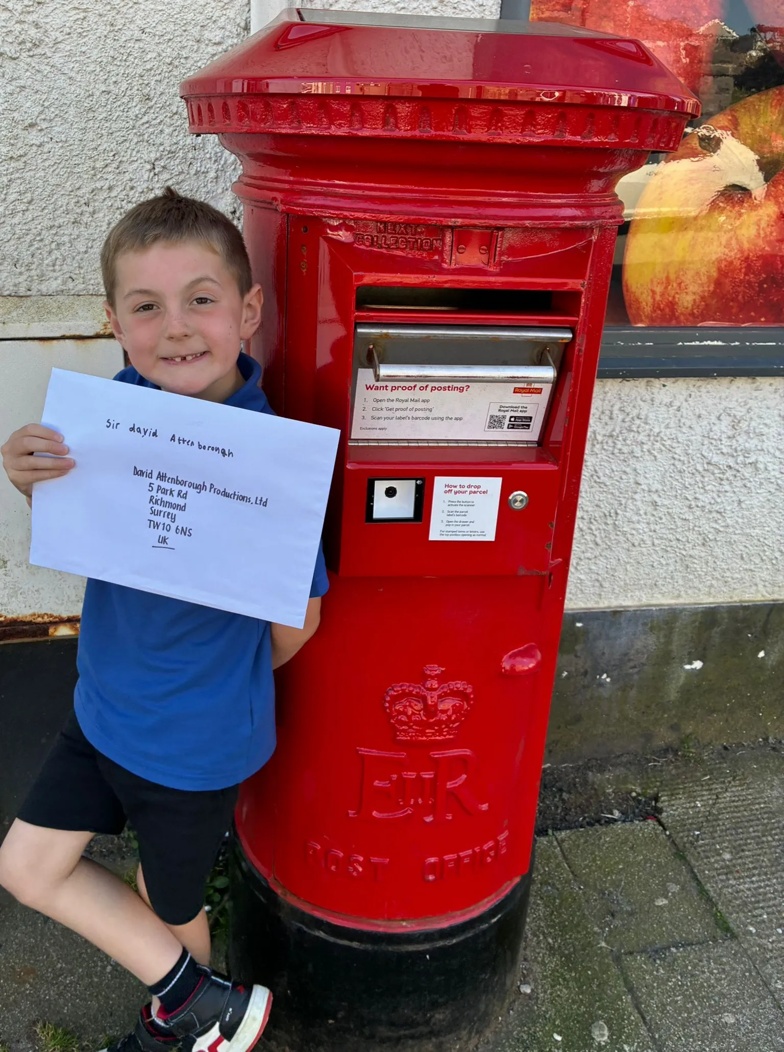 Samantha Evans-Browning Max stands leaning against a bright red post box. he holds a large A3 sized white letter in front of his chest and smiles at the camera. 