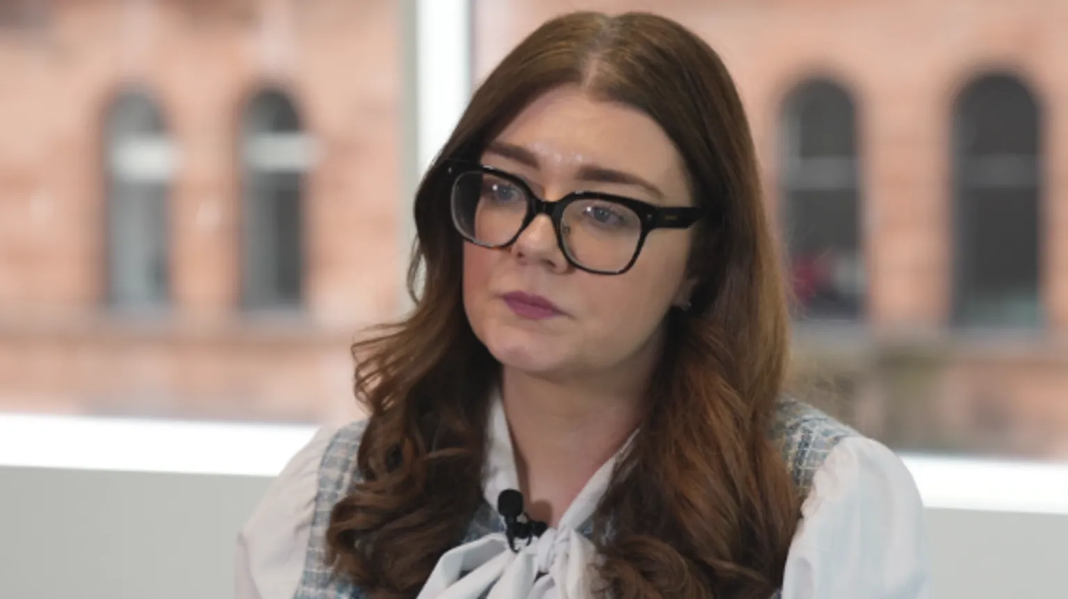 Jennifer Lilly is a young woman with long red-brown hair, large plastic spectacles and a white blouse. She is in an office with a red sandstone building visible through the window.