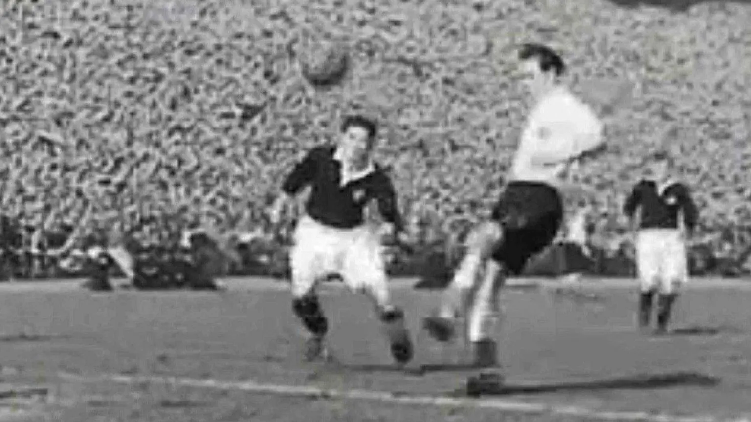 A black‑and‑white action shot shows two footballers contesting a bouncing ball near the penalty area. One player is lifting a leg to strike while another moves in to challenge, with additional players further back. A tightly packed crowd fills the stadium stands in the background, highlighting the scale and atmosphere of the match.