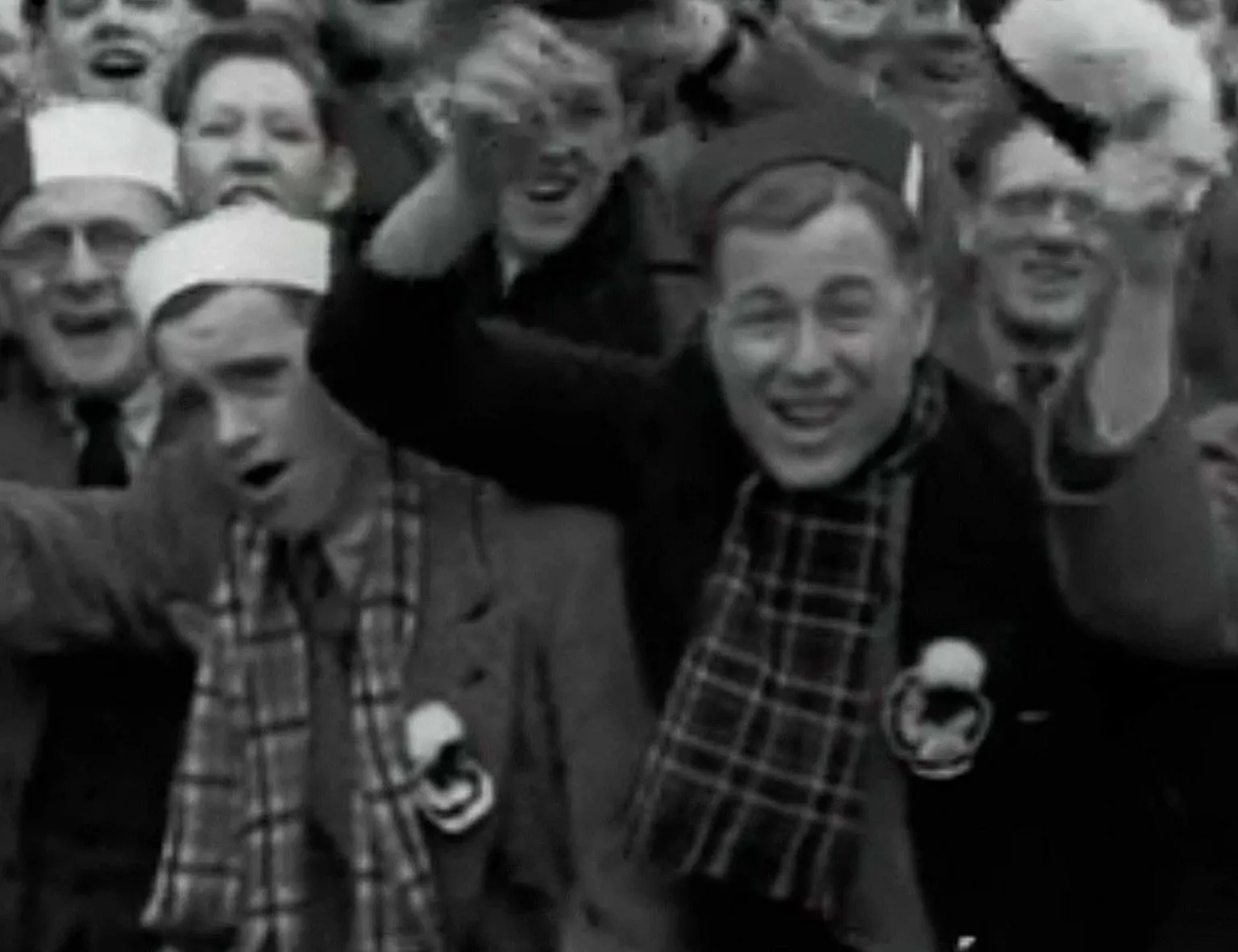 A tightly packed group of spectators fills the frame, gathered closely together in a stadium stand. Several individuals in the foreground wear scarves and hats, with raised arms and open mouths suggesting active cheering or chanting. The black‑and‑white image captures the dense crowd and energetic match‑day atmosphere.