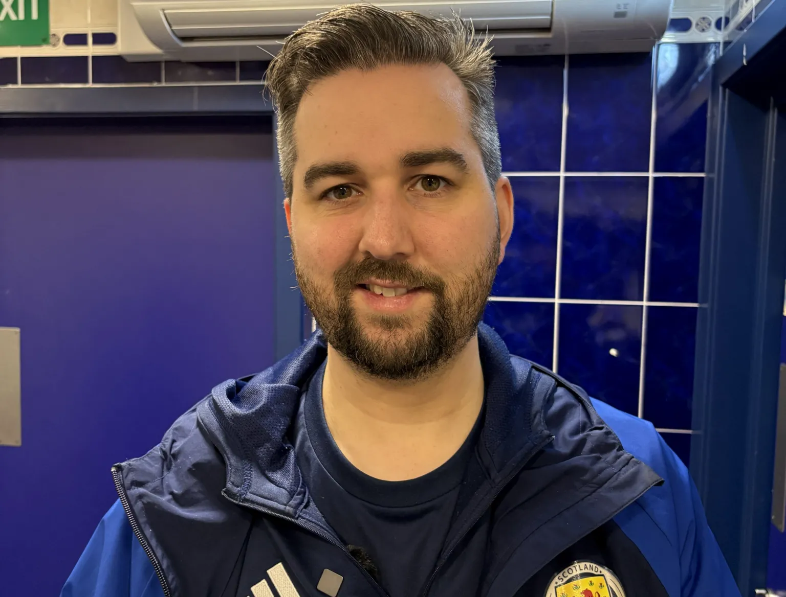 Andy Kerr stands in one of the Hampden dressing rooms, against blue tiled walls, with a visible air-conditioning unit above. He is wearing a dark jacket with a Scotland badge. 