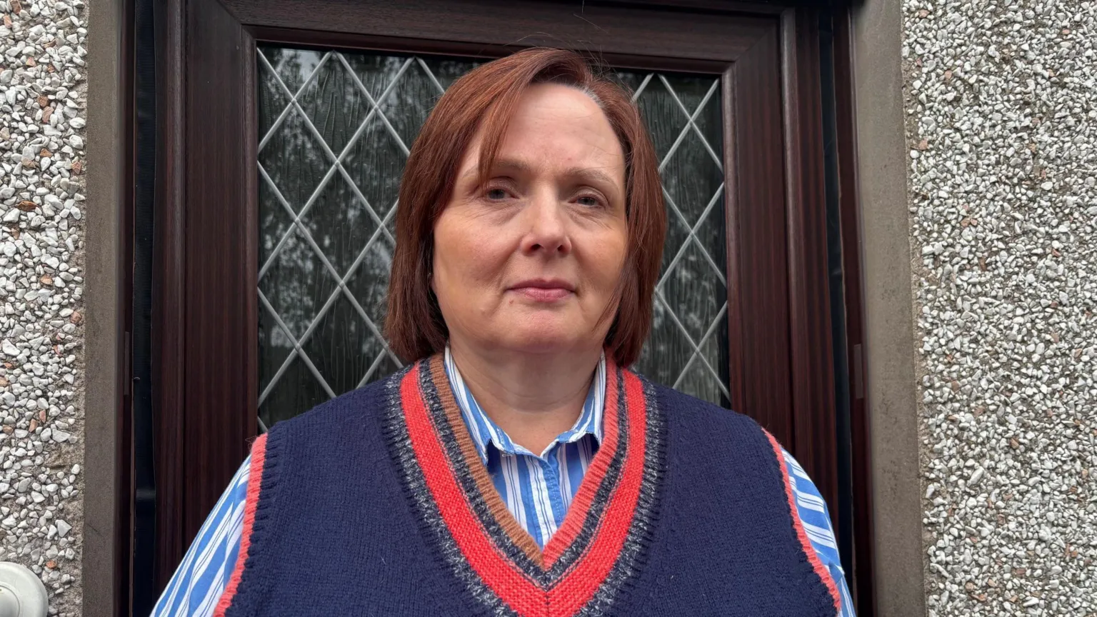 A middle-aged woman is standing in front of the door of a house. The door is dark brown wood effect, with a leaded style glass panel. She has chin length auburn hair and is wearing a blue knitted vest top over a blue and white striped shirt. 