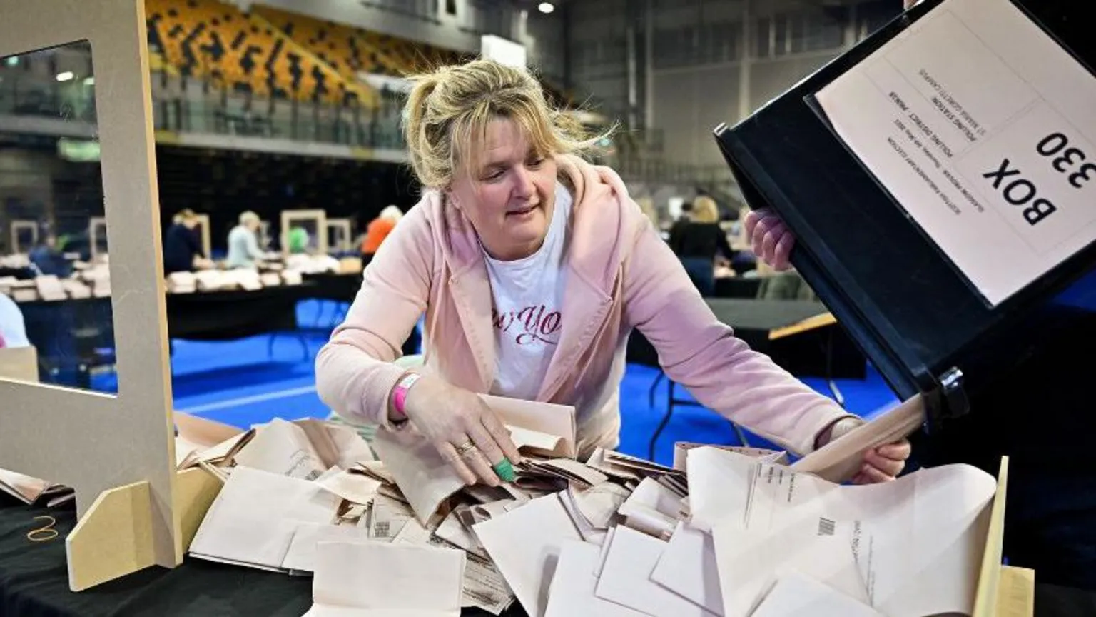  A woman leans over a table where a large pile of ballot papers are being pourer from a black box. The woman, wearing a pink hooded jumper, is in a large hall with counting desks and an empty stand of orange seats in the background.