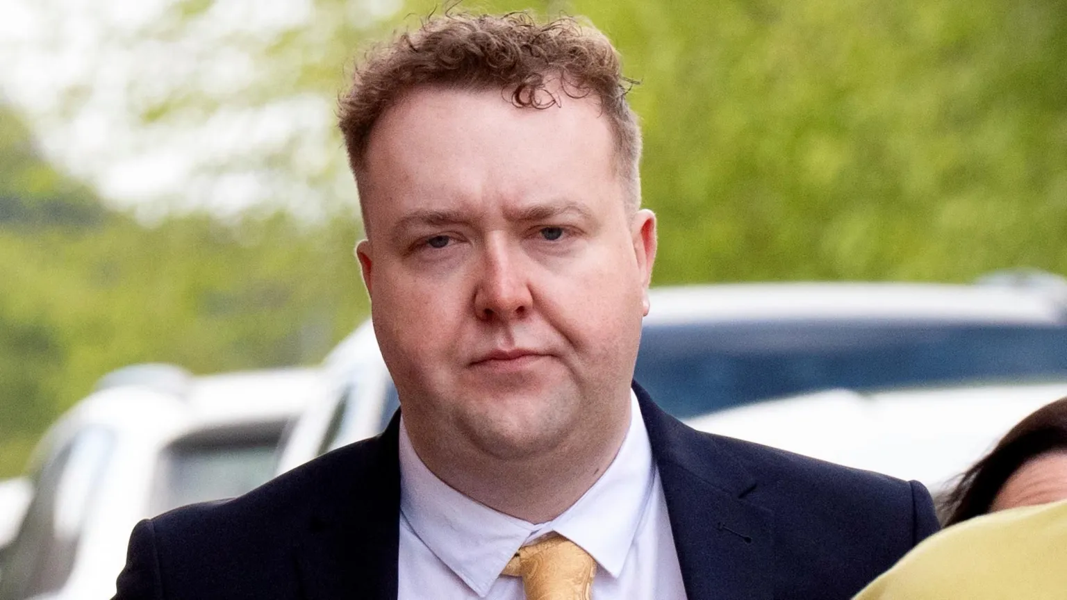  Jordan Linden arrives at court. He has short brown hair and is wearing a dark blue suit, light pink shirt and a yellow tie.