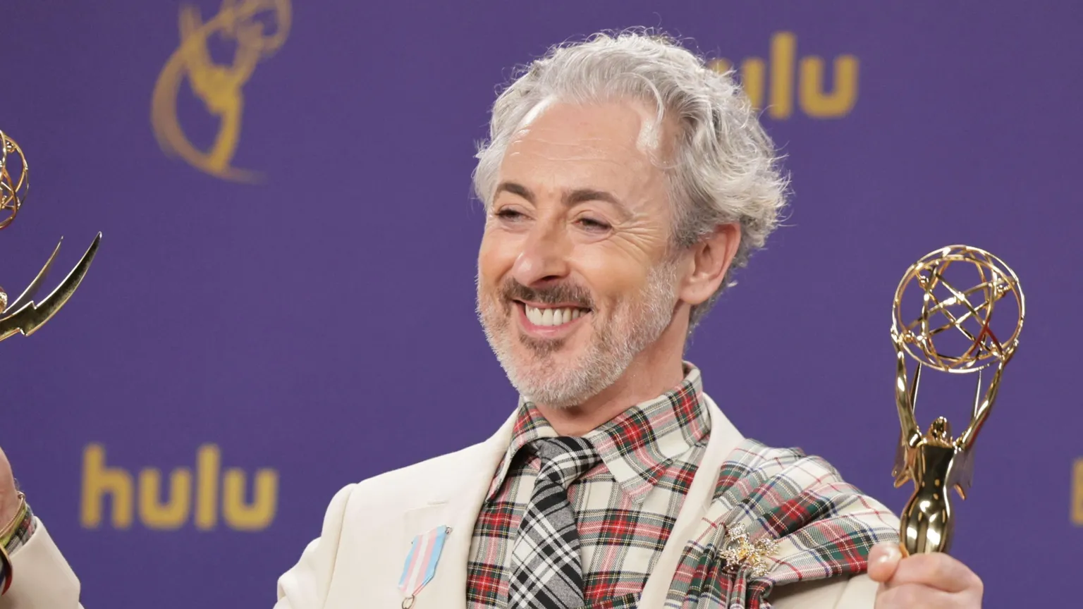 EPA Alan Cumming wearing a cream jacket with a tartan shirt and tie. He is smiling at the camera holding two trophies.