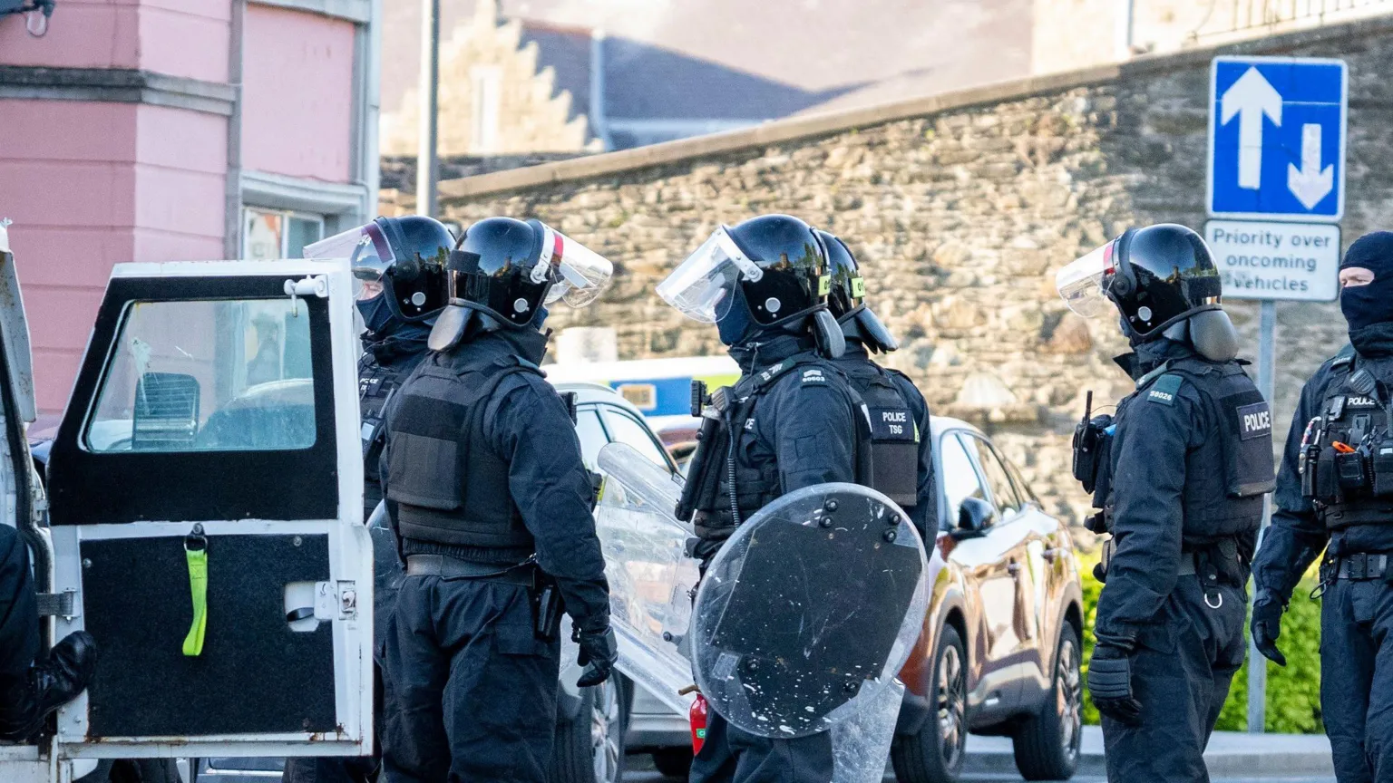 Aodhán Roberts A number of police officers in black uniform and wearing helmets hold shields as they stand in a group. The open door of a police vehicle is to the left of the image