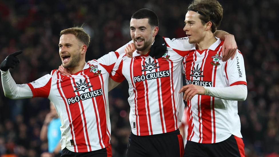 Finn Azaz, wearing Southampton's red and white striped home shirt, smiles as he celebrates scoring a goal against QPR. His team-mate Leo Scienza stands with his arm around Azaz with fist pumped. Caspar Jander stands with his arm around Azaz on the other side.