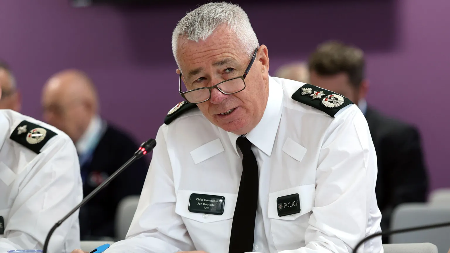  PSNI Chief Constable Jon Boutcher, sitting at a table with a microphone in front of him. He has short grey hair and is wearing glasses, a white shirt and black tie. 