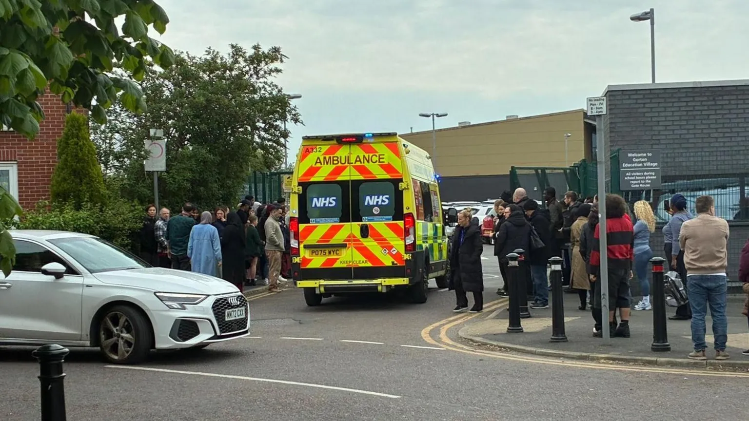 An ambulance pulls at the school. A number of people are waiting outside the school. 