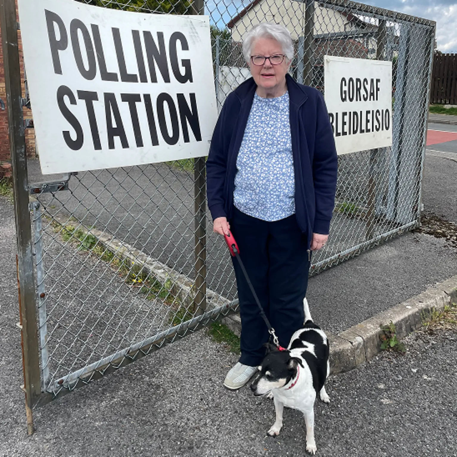 Kay Sebern outside a polling station with her Jack Russel Sasha - Kay has a navy blue top and trousers on with a light blue shirt underneath. Sasha is black and white and being held on a retractable leash. The English and Welsh polling station signs are mounted to the chainlink fence behind her 