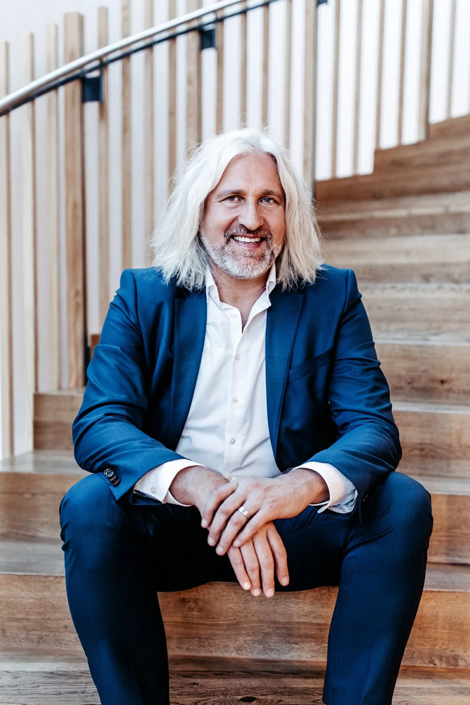 Christian Ege Head shot of Christian Ege with shoulder-length grey hair and beard sitting on some wooden steps. He is wearing a blue suit with a white shirt with the top buttons unopened.