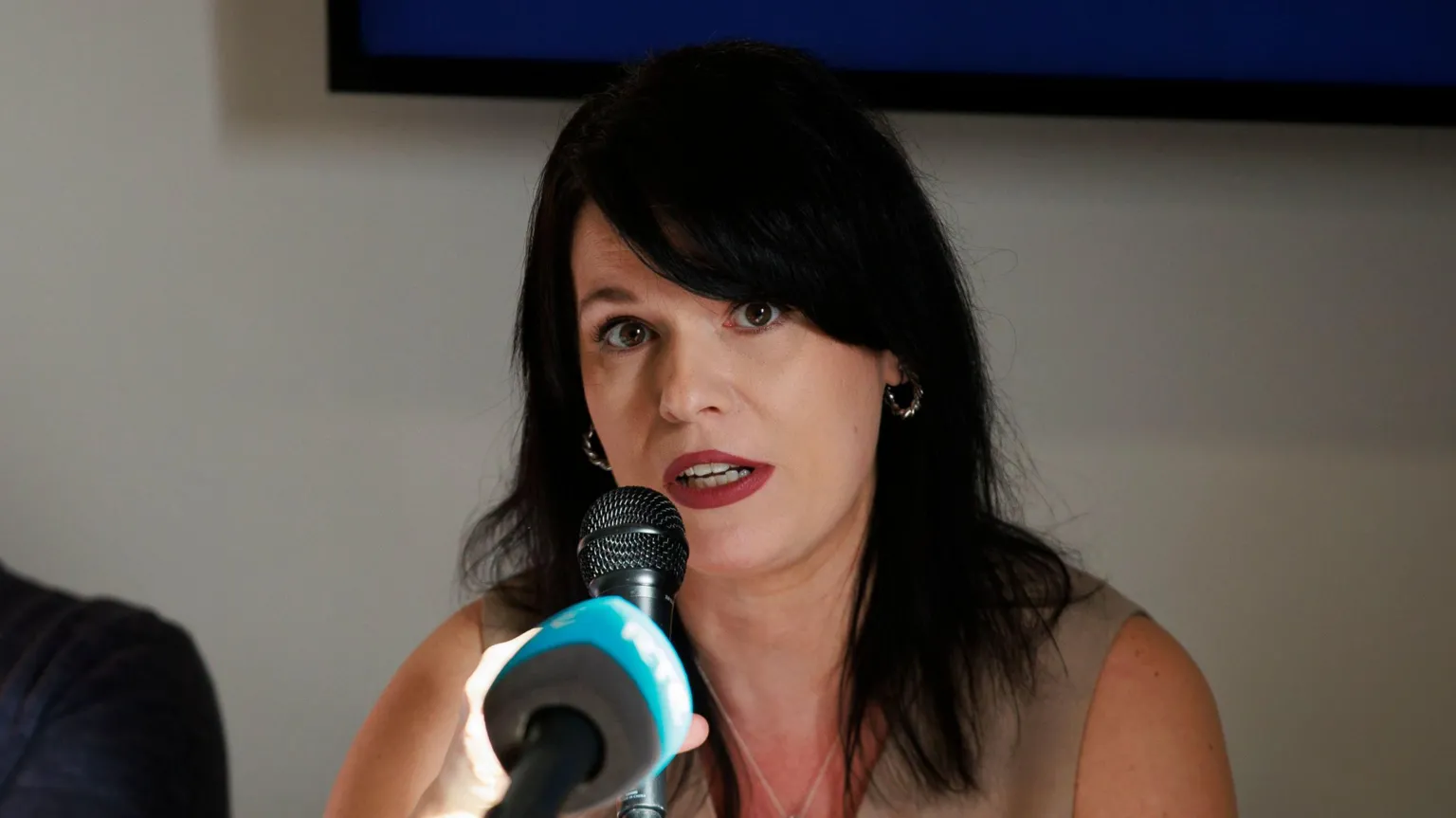  A woman, sitting in front of a blue microphone. She has a sleeveless cream coloured top on and has long black hair. 