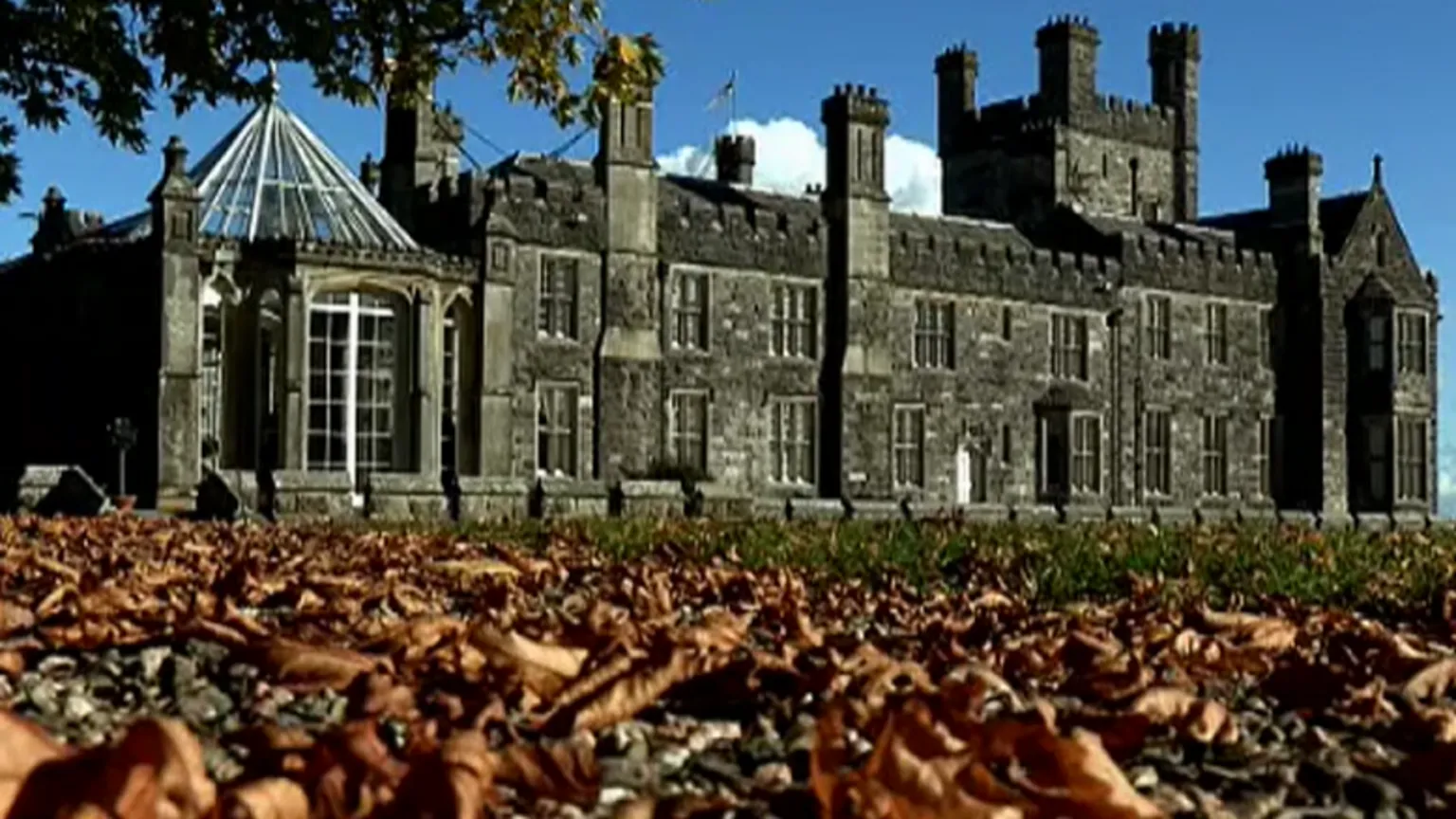 Crom Castle is a large grey stone building. It has many windows and a glass conservatory. The picture has been taken in autumn as brown leaves lie on the ground in front of the castle. 
