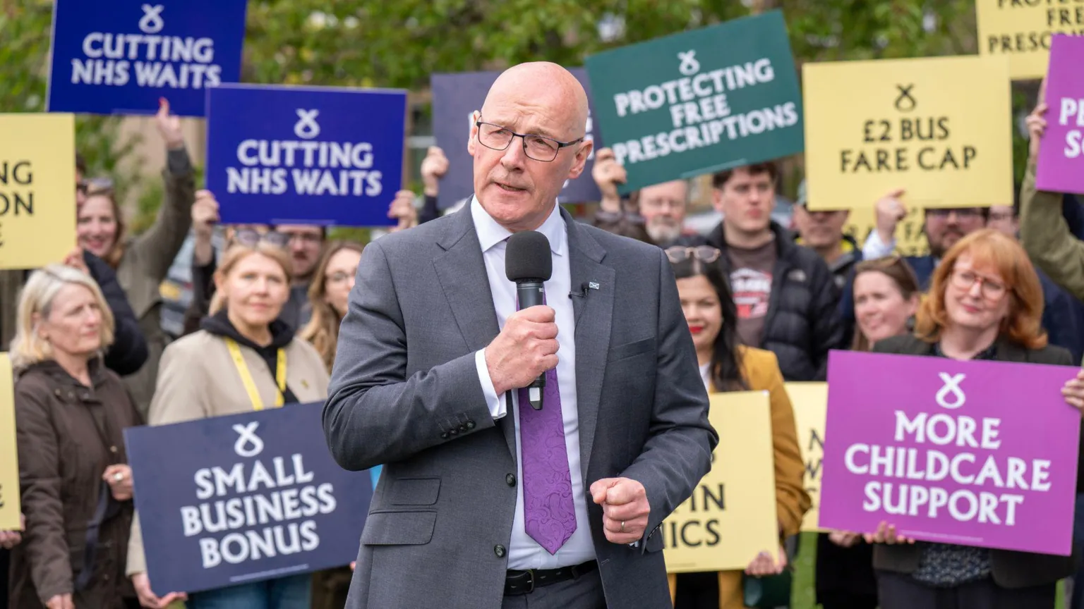  John Swinney standing in front of a group of MSPs and supporters holding signs in support of the SNP.