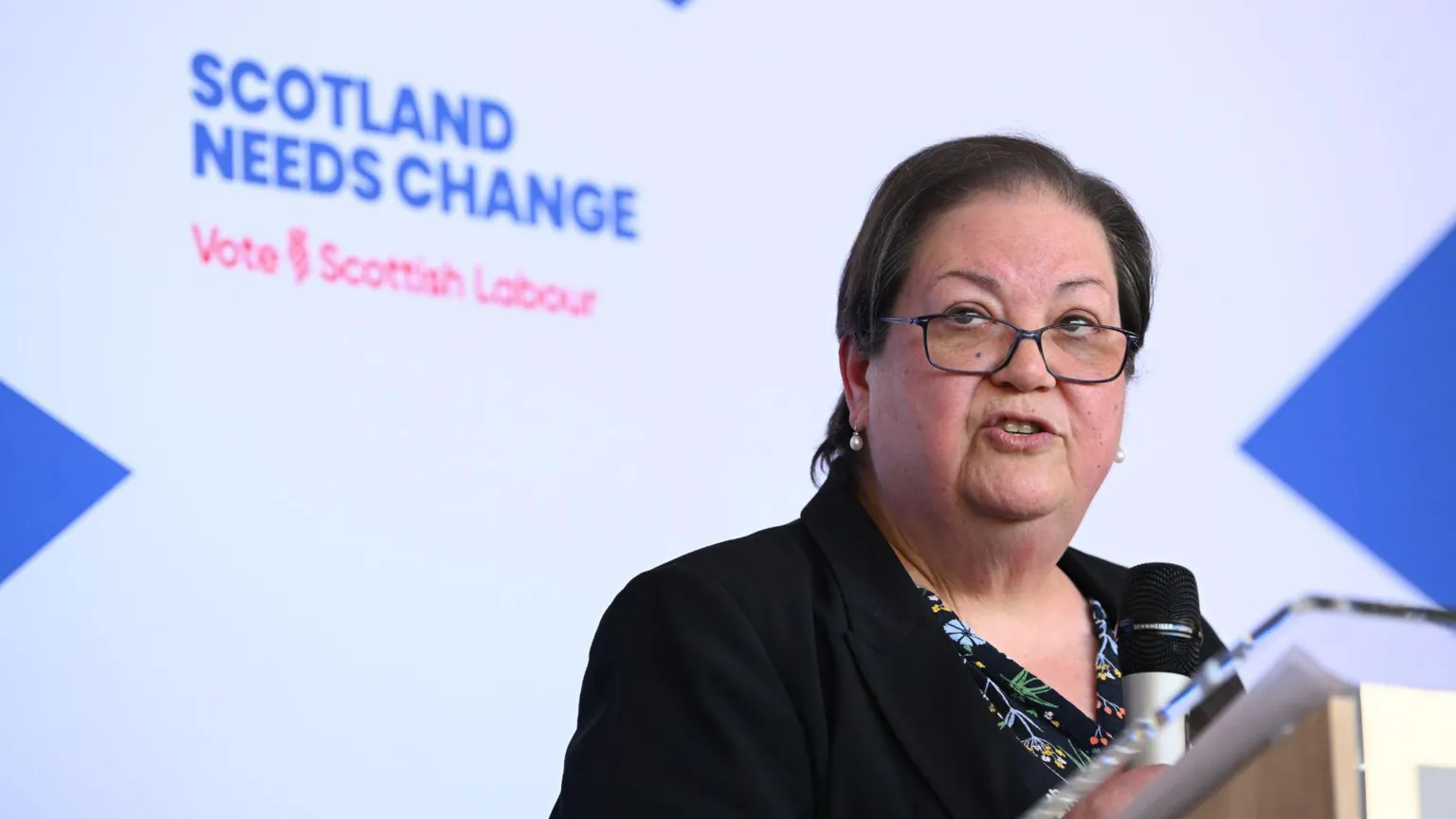  Jackie Baillie standing at a podium in front of a large saltire background with a Scottish Labour election message on it.