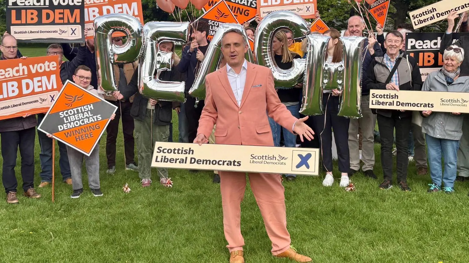  Alex Cole-Hamilton holding a Scottish Liberal Democrats banner wearing a peach suit and standing in front of supporters holding placards