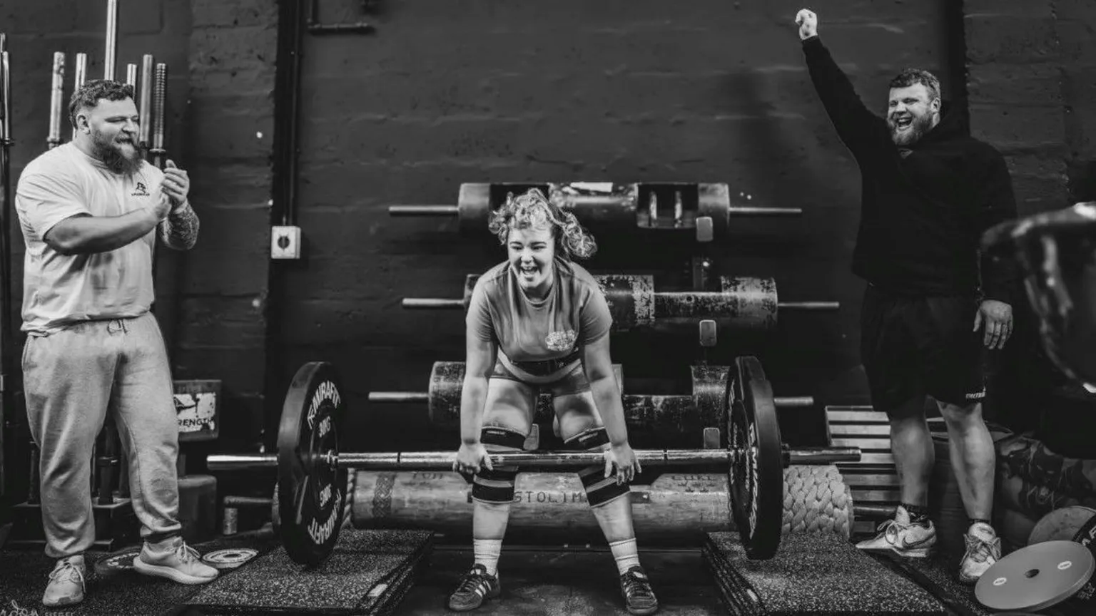 Gordon Siegal A black-and-white shot of lauri, laughing as she starts to lift a barbell with huge weights on each end. Her hair is in a curly ponytail and the Stoltman brothers, both strong-looking men with beards, are cheering her on from the sides.