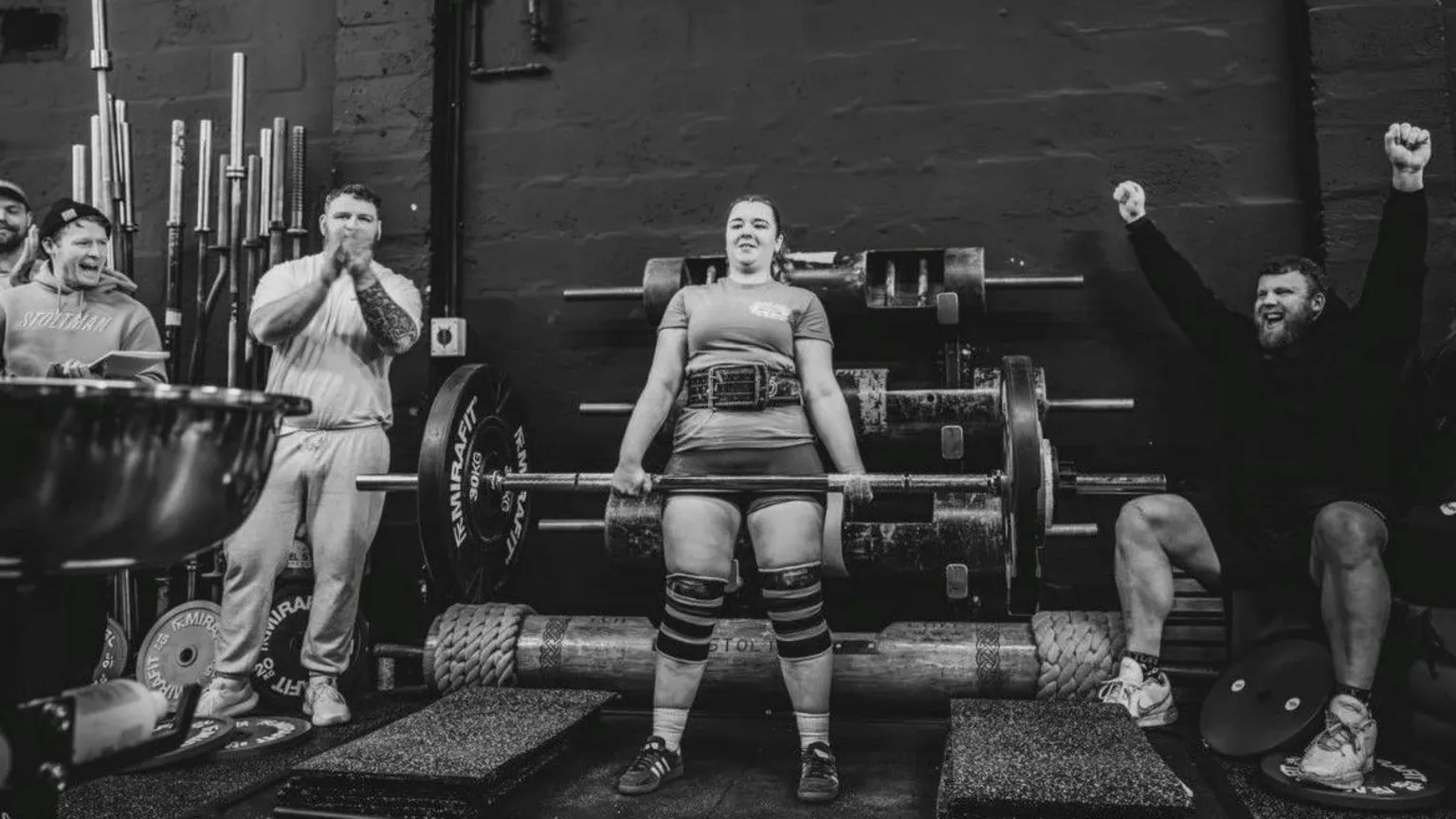 Gordon Siegal A black‑and‑white gym scene shows Lauri in the centre performing a heavy deadlift on a platform surrounded by weights. Several individuals, including the Stoltman brothers, stand nearby clapping and raising their arms, positioned along the sides of the lifting area. Barbells, plates, and gym equipment line the background against a dark wall.