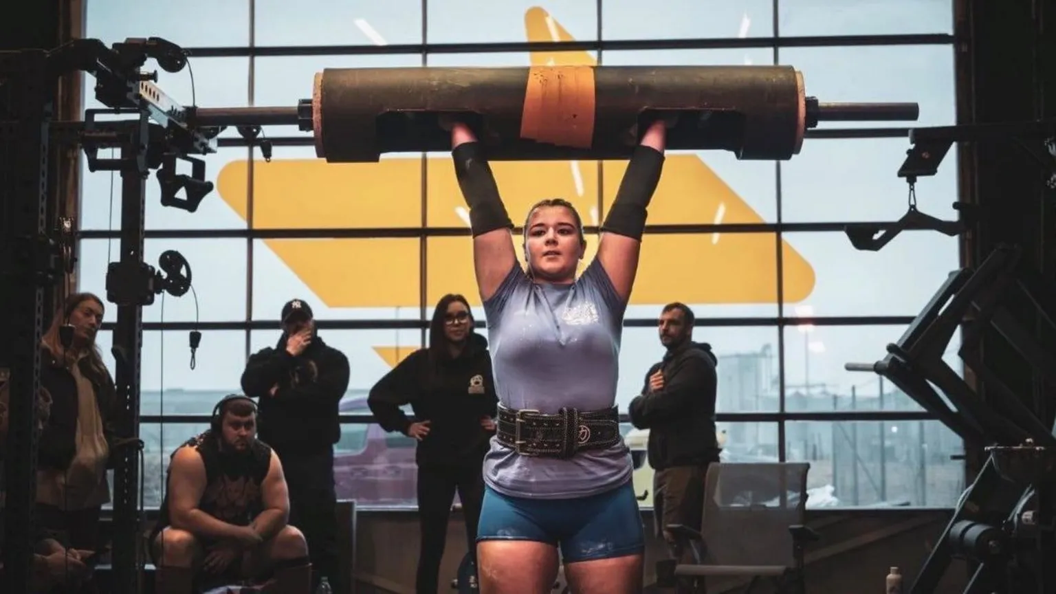 Gordon Siegal Lauri stands in a gym, lifting a large cylindrical bar overhead with both arms extended. Several onlookers and gym equipment surround the platform, with a large window behind letting in muted daylight. There are belts, racks, and machines visible.