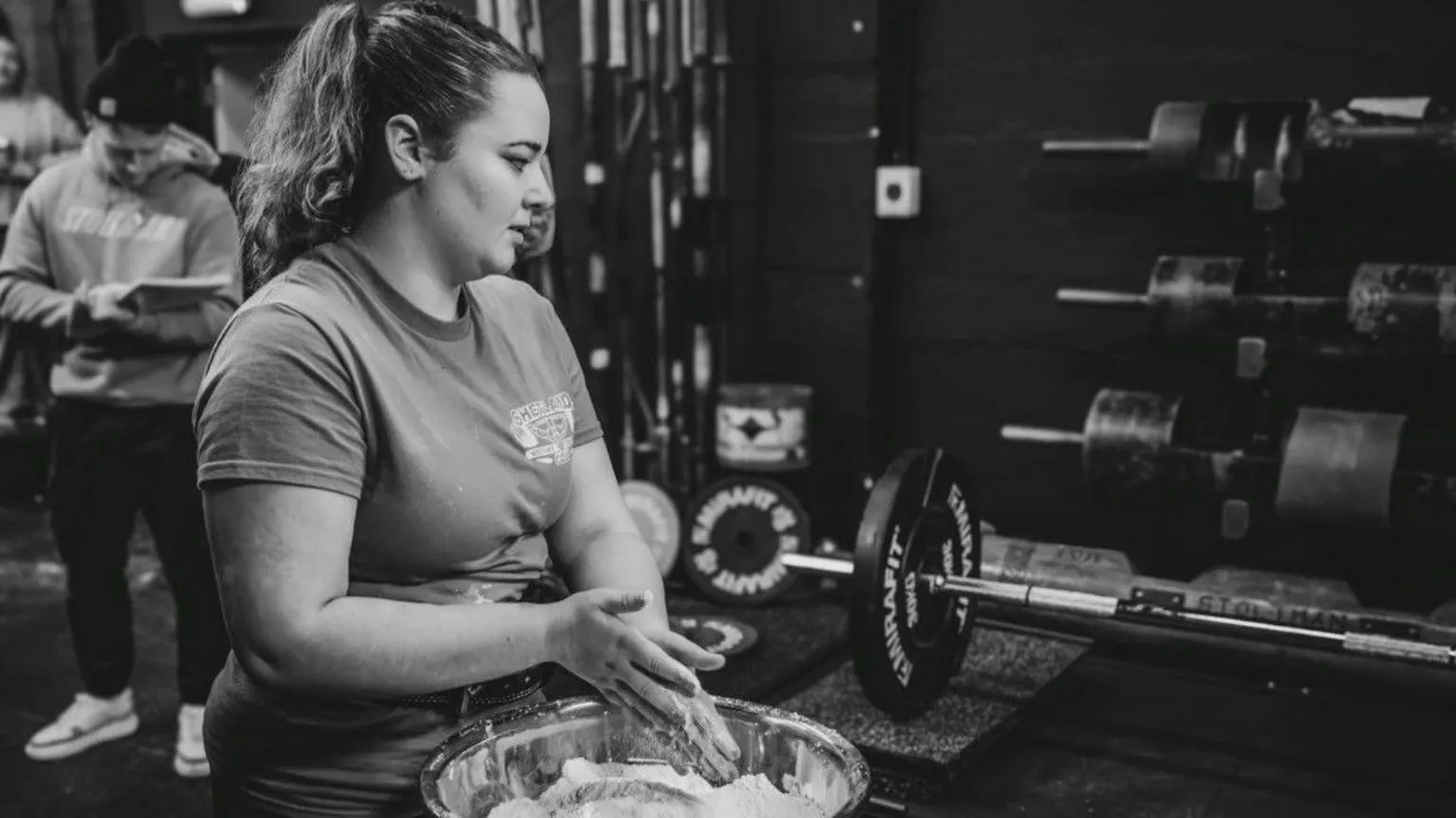Gordon Siegal A black‑and‑white image shows Lauri standing beside a large bowl filled with chalk, rubbing hands in preparation for lifting. A loaded barbell rests on a platform nearby, with weight plates and racks visible against a dark wall in the background. Another individual stands behind holding a notepad, while various strength‑training equipment surrounds the area.