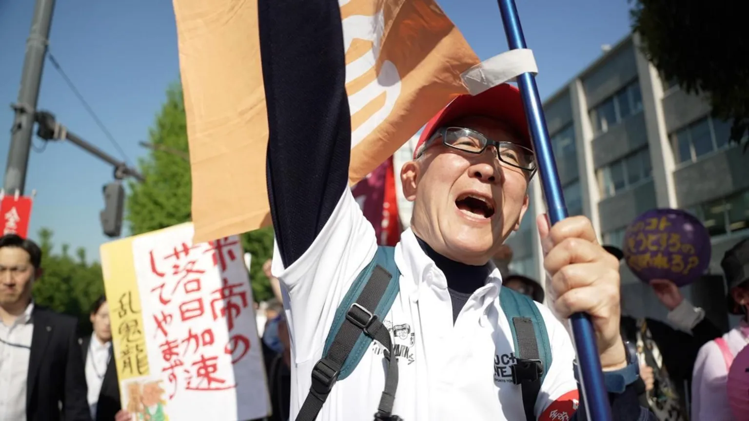 : Jiro Akiba A man in a backpack, glasses and a red cap punches a hand in the air while holding a flag