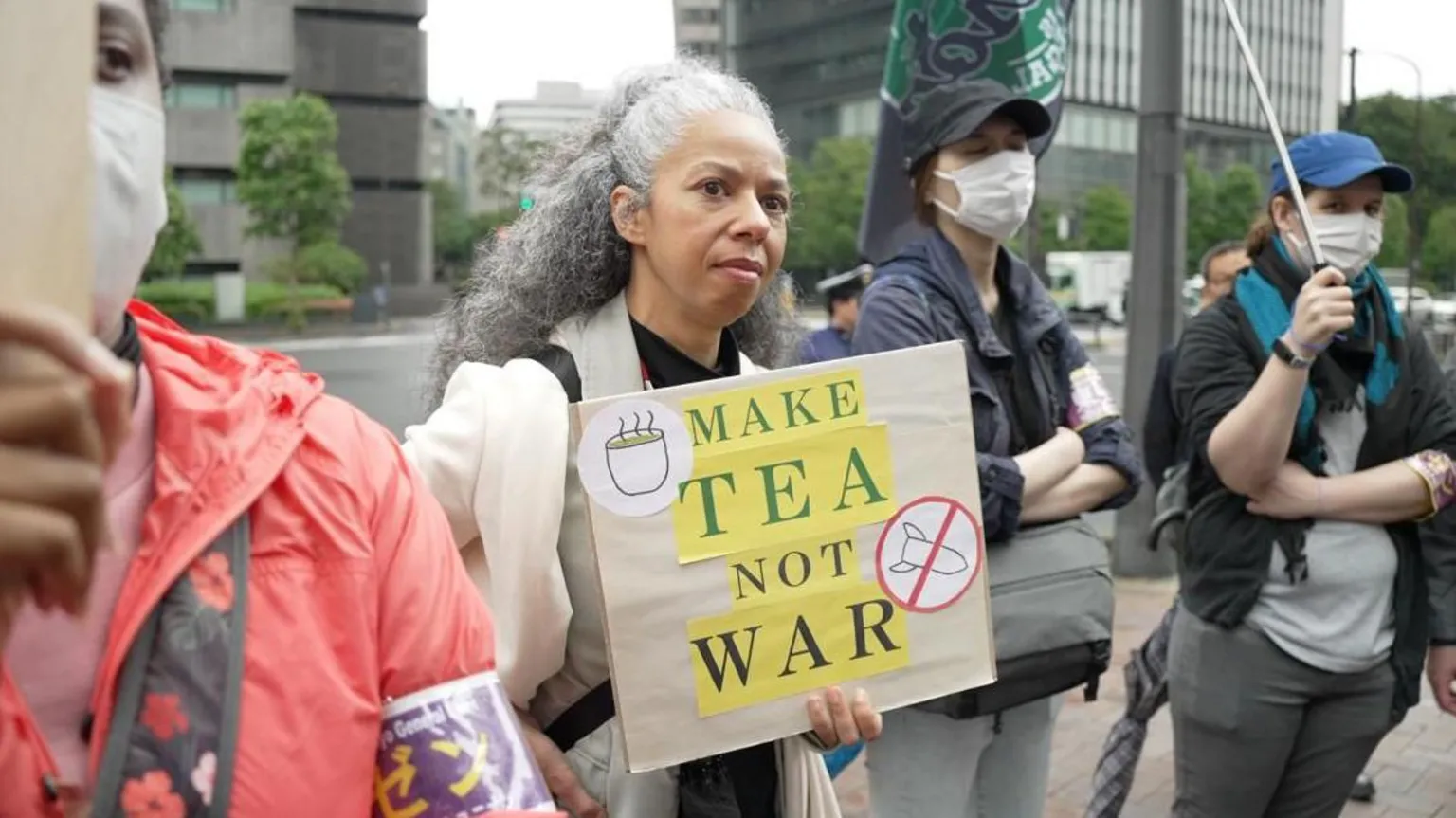 : Jiro Akiba A woman with grey hair holds a sign that says 'Make tea not war'