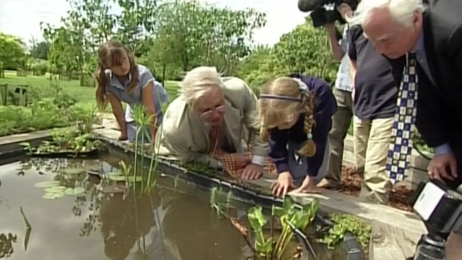 Sir David Attenborough is pictured looking into a pond with two schoolchildren. He is wearing a white suit. To the right of the image another older man wearing a dark suit leans over and peers into the pond as well. 