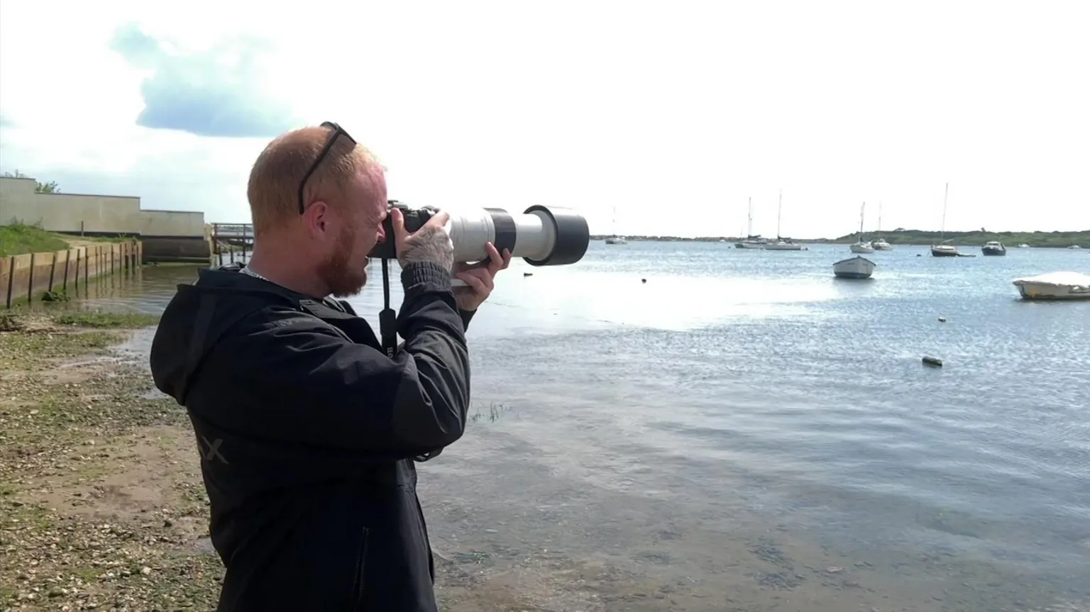 Lewis Slavin stood on the side of the water where there are boats floating in the distance. He is wearing all black and has a large camera that he is taking photographs with.