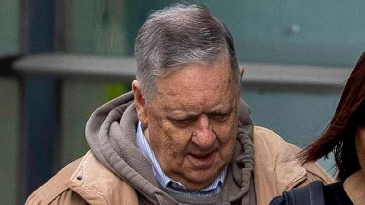  Lloyd-Lavery has livery skin, spotted with marks from ageing. His relatively full head of hair is grey and combed to the side. He wears a sandy jacket, grey hoodie and stiped shirt. 