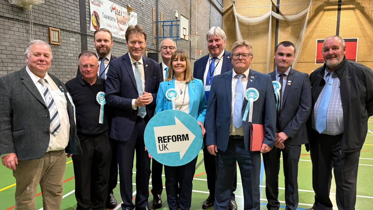 A group of nine men and one woman are standing in a sports hall. They are mostly wearing suits. The woman is standing in the centre and is holding a light blue Reform UK sign.