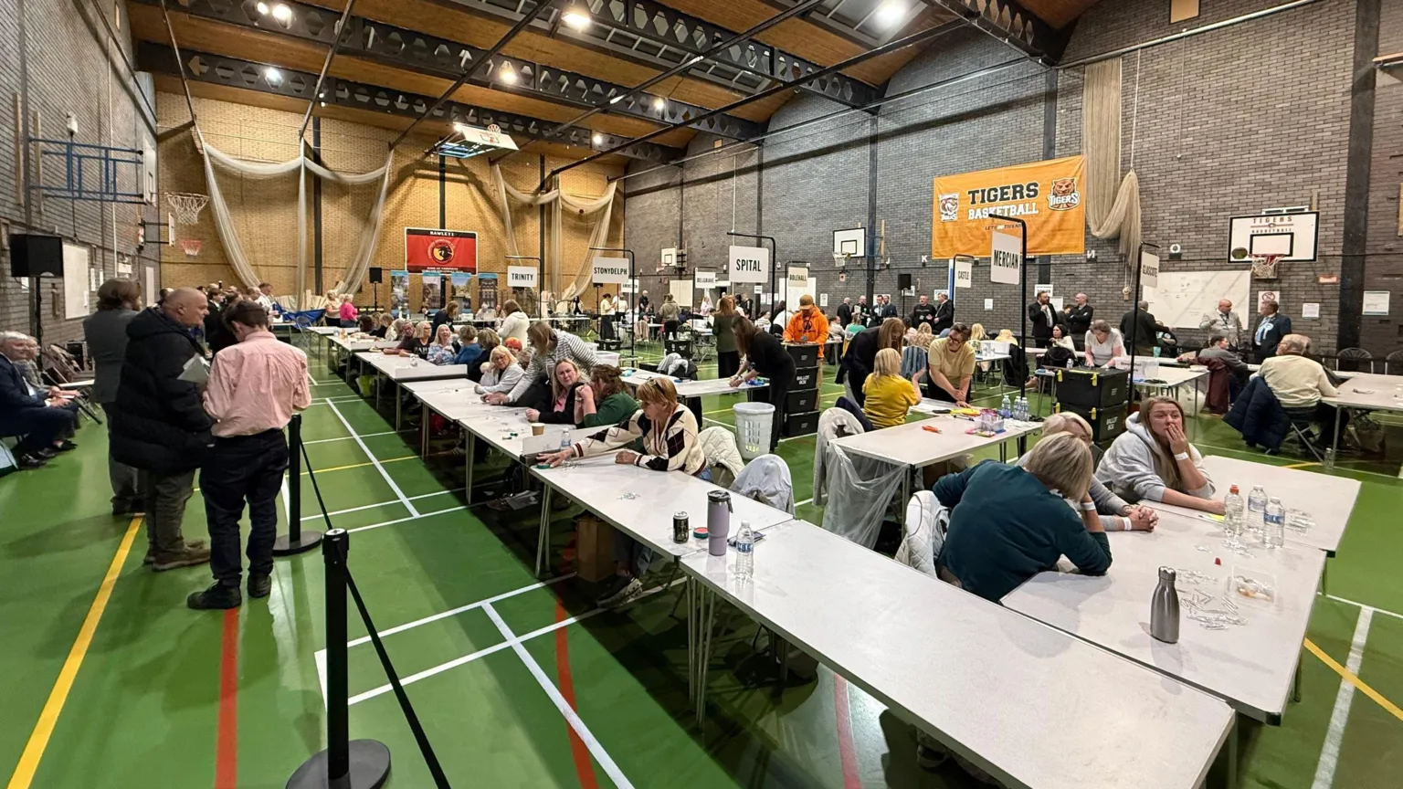 A large sports hall with a green floor. There are tables set out with people sat at them preparing to count ballots. There are others stood around talking to one another. On the wall is an orange banner for a basketball team and there are various drinks bottles on the tables.