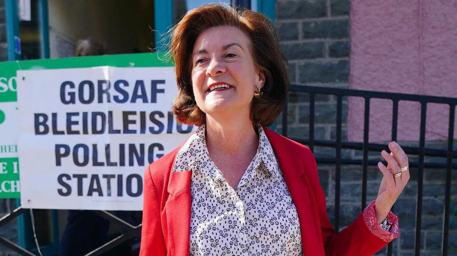  Eluned Morgan stood in front of a sign that says polling station in English and Welsh. She is wearing a red jacket, a white and black shirt with patterns and is smiling to the camera.