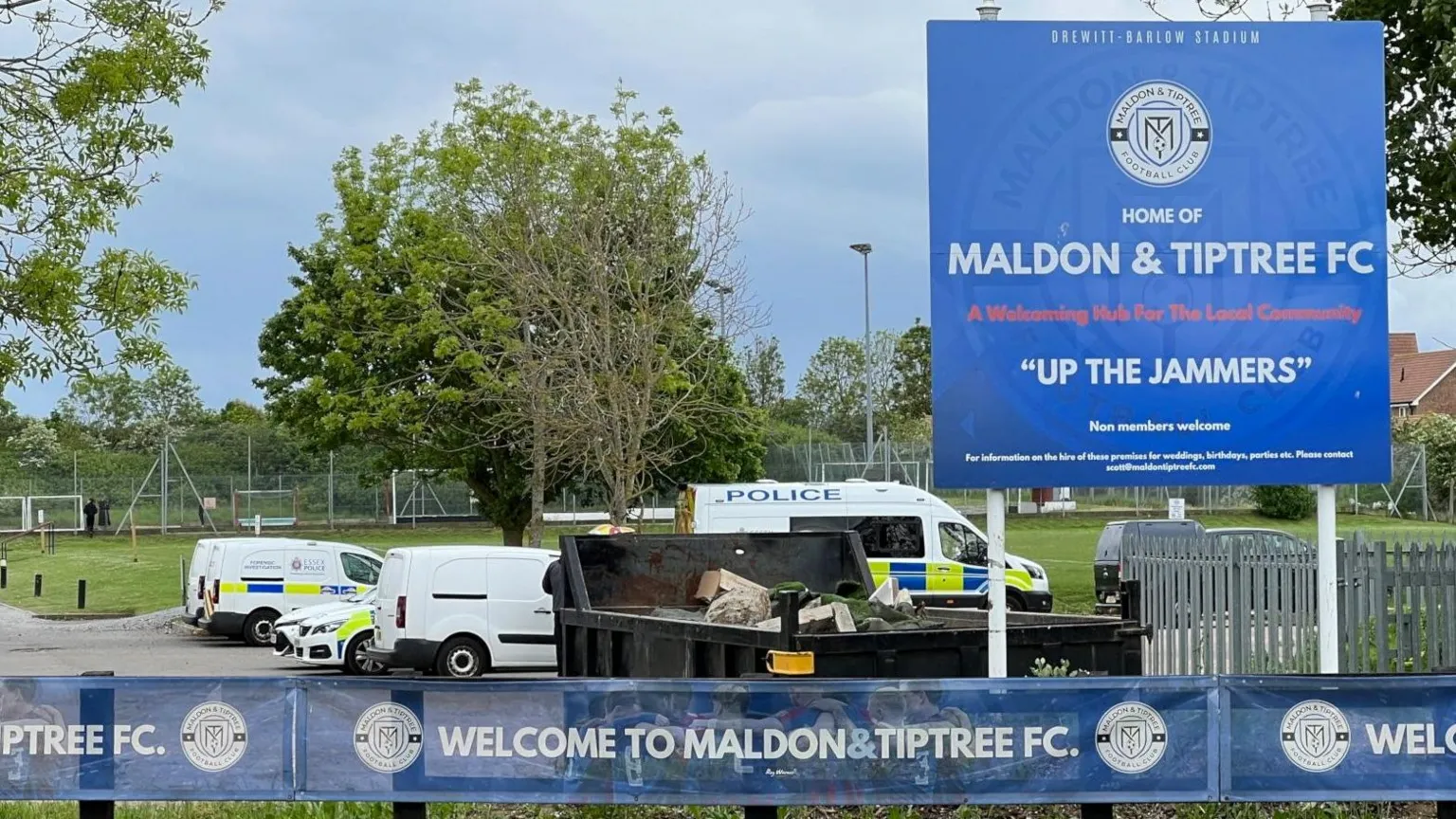 Stuart Woodward/BBC A blue sign reads Home of Maldon and Tiptree FC . Police cars and vans are parked up in the background.