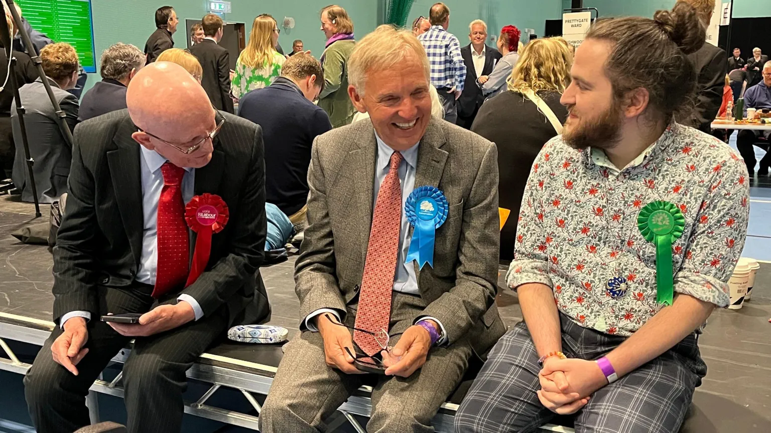 Charlotte Rose/BBC Three male candidates (left to right) with red Labour, blue Conservative and Green rosettes sit on a stage in a busy counting hall.