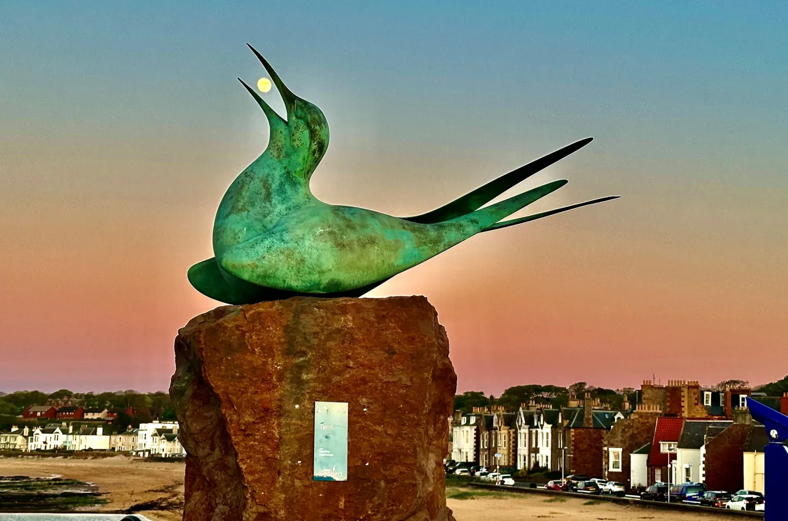 Pat Christie A statue of an arctic tern. It has its head raised upwards, and mouth open, so it appears to be eating the full moon visible between the gap. 