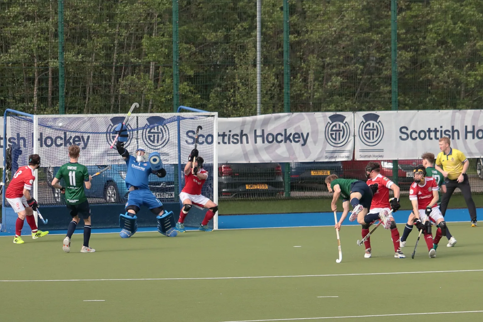 Gordon Baxter A goal being scored in a hockey match. The scoring team are wearing green shirts and black shorts. One of their team is airborne as he watches his shot go in. The other team are in red shirts and white shorts with their netminder in all blue. A referee in a yellow shirt watches on.