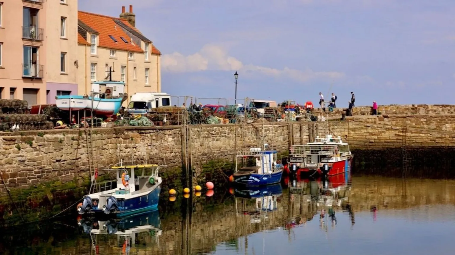 Graham Christie A row of brightly painted three fishing boats at a harbour. Several people can be seen walking around in the background of the harbour.