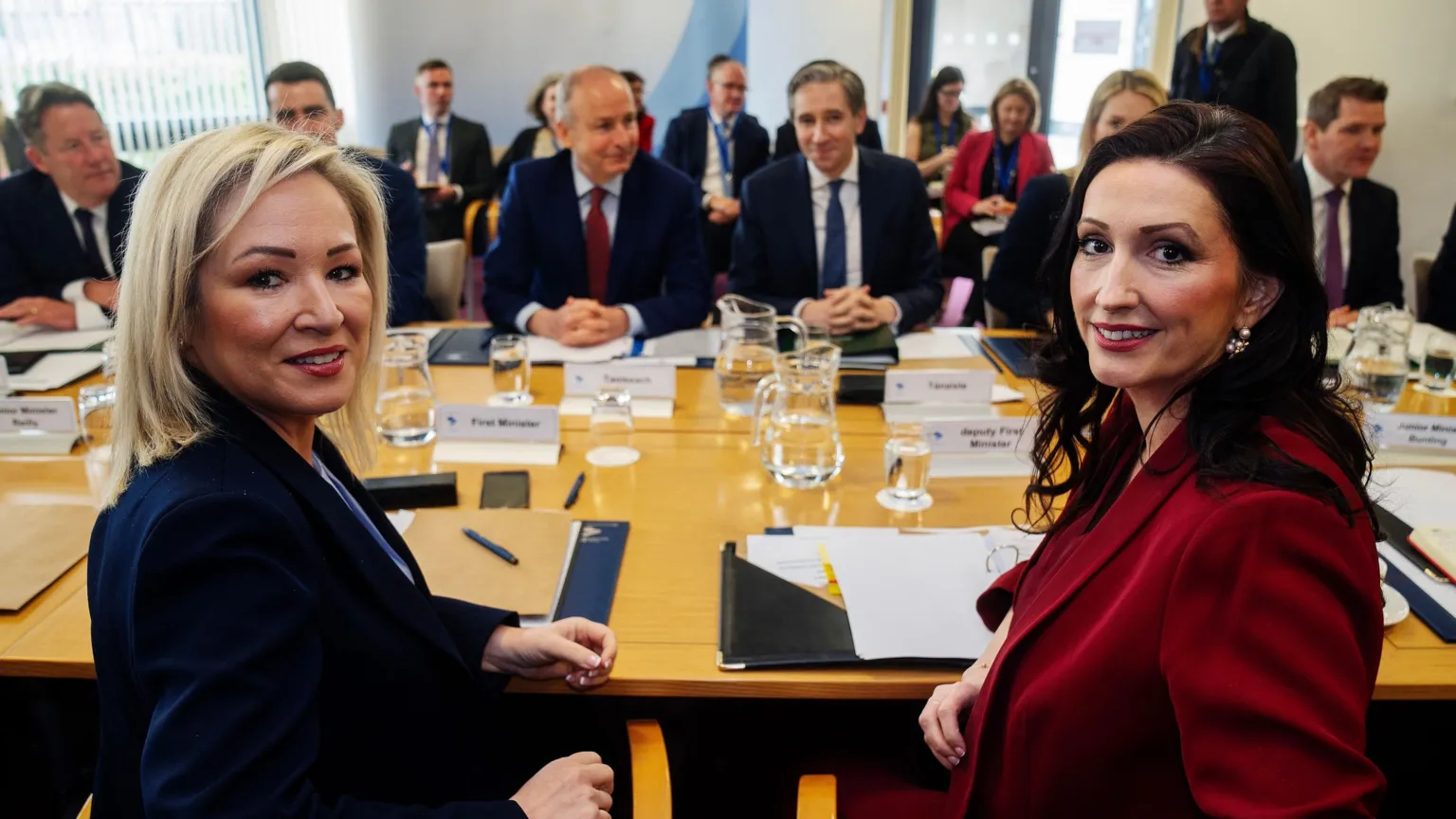 Brian Lawless/PA Wire Michelle O'Neill and Emma Little-Pengelly sitting a large round conference table, turning towards the camera and smiling. Sitting across from them is Micheál Martin and Simon Harris and other attendees. There are folders, jugs of water and name tags on the table.