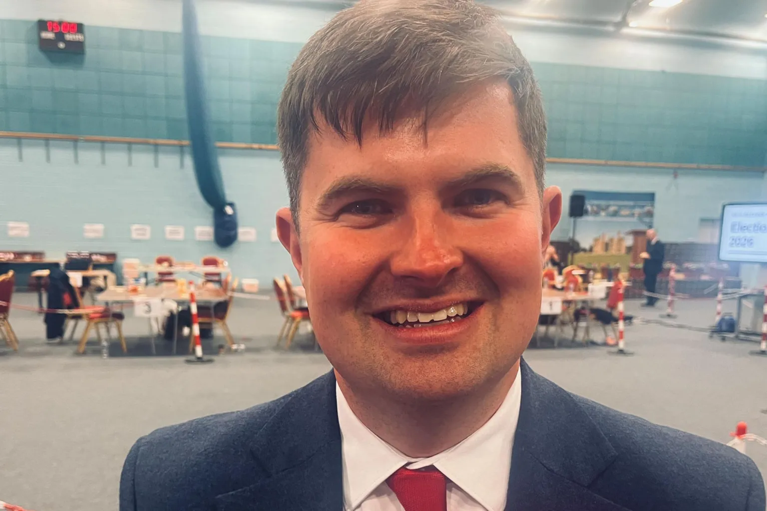 Donald MacKinnon has dark hair and is wearing a dark suit over a white short and red tie. He is wearing a red rosette.