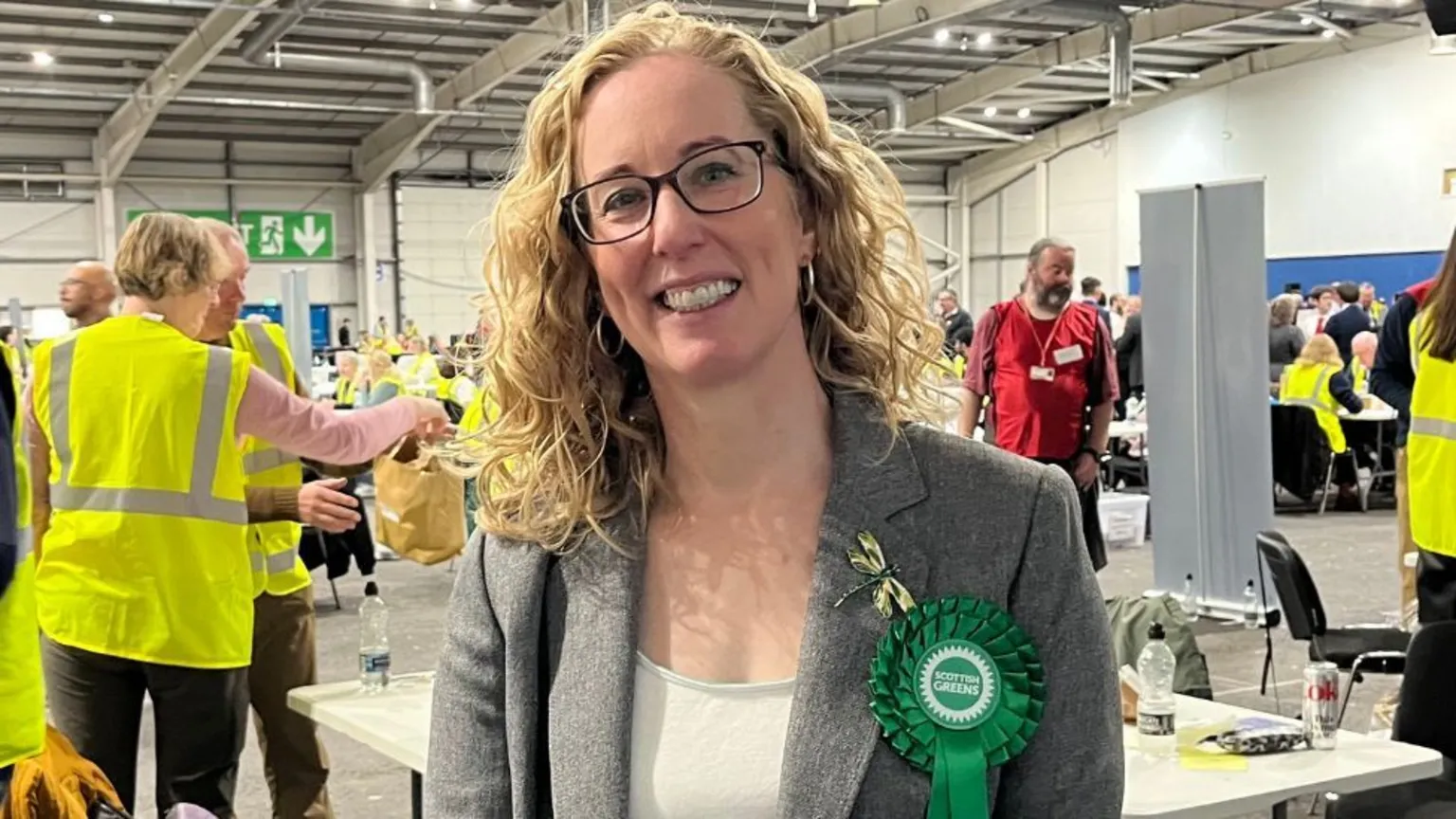 Lorna Slater, who has curly blonde hair, smiles in a counting centre. She is wearing a Green rosette 
