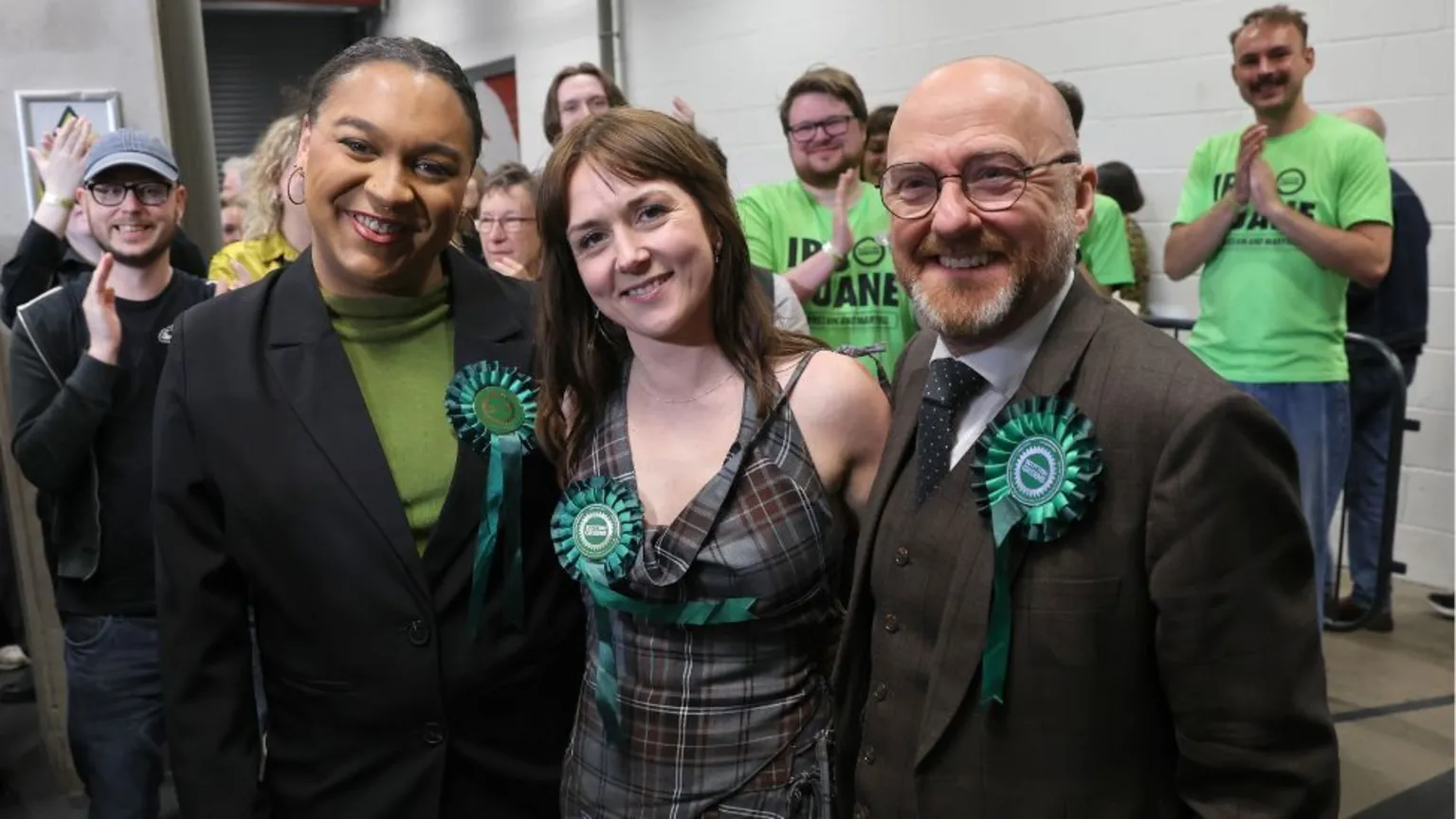  Three smiling candidates with Green party rosettes pose in front of clapping activists 