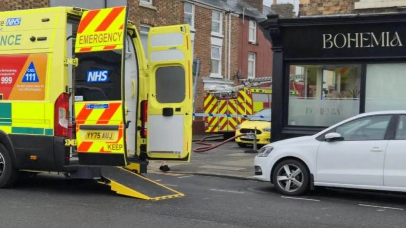 Lou Lambert The image shows an emergency response scene on a residential street. A shopfront with the sign BOHEMIA is visible on the right side. A bright yellow NHS ambulance is parked on the street, with the rear doors open.
